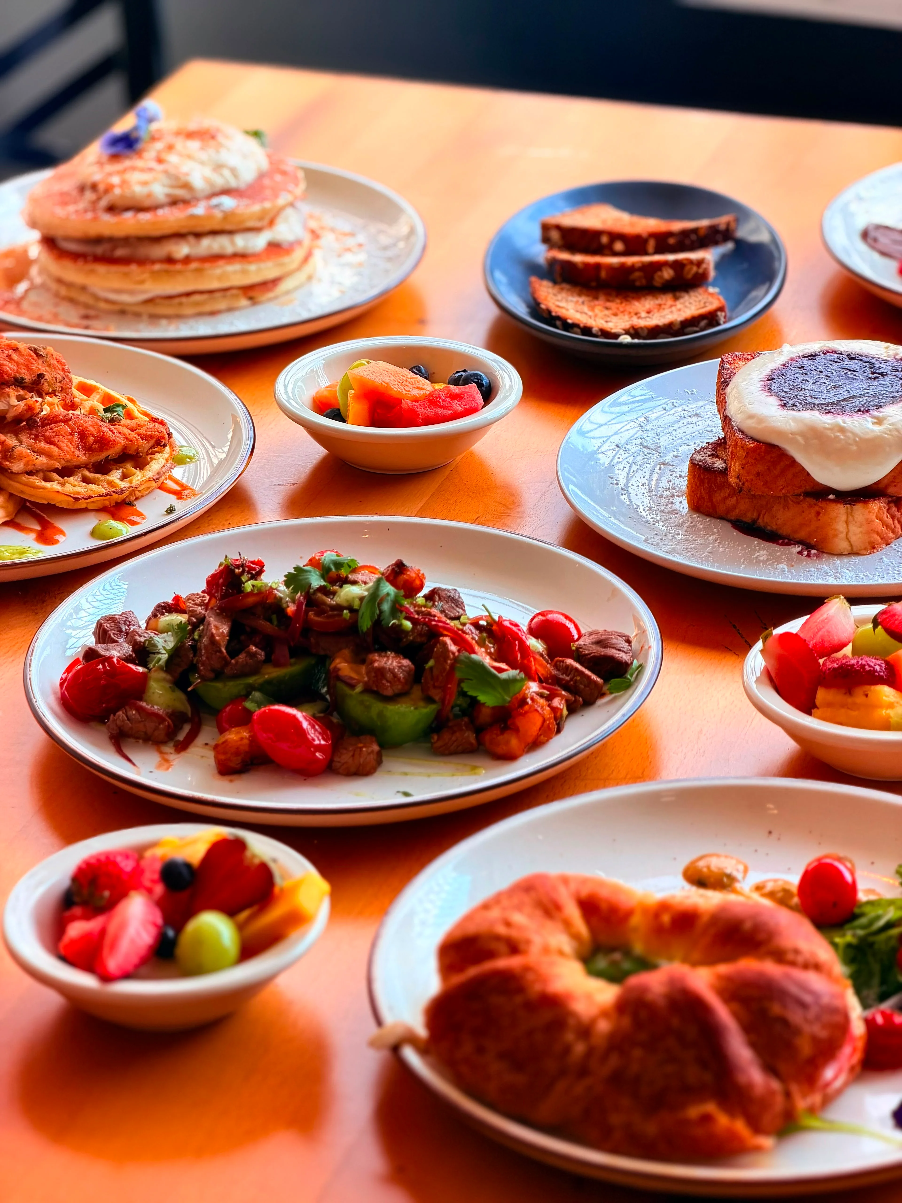 An array of brunch items on a wooden table, including pancakes, colorful salads, and different types of bread.