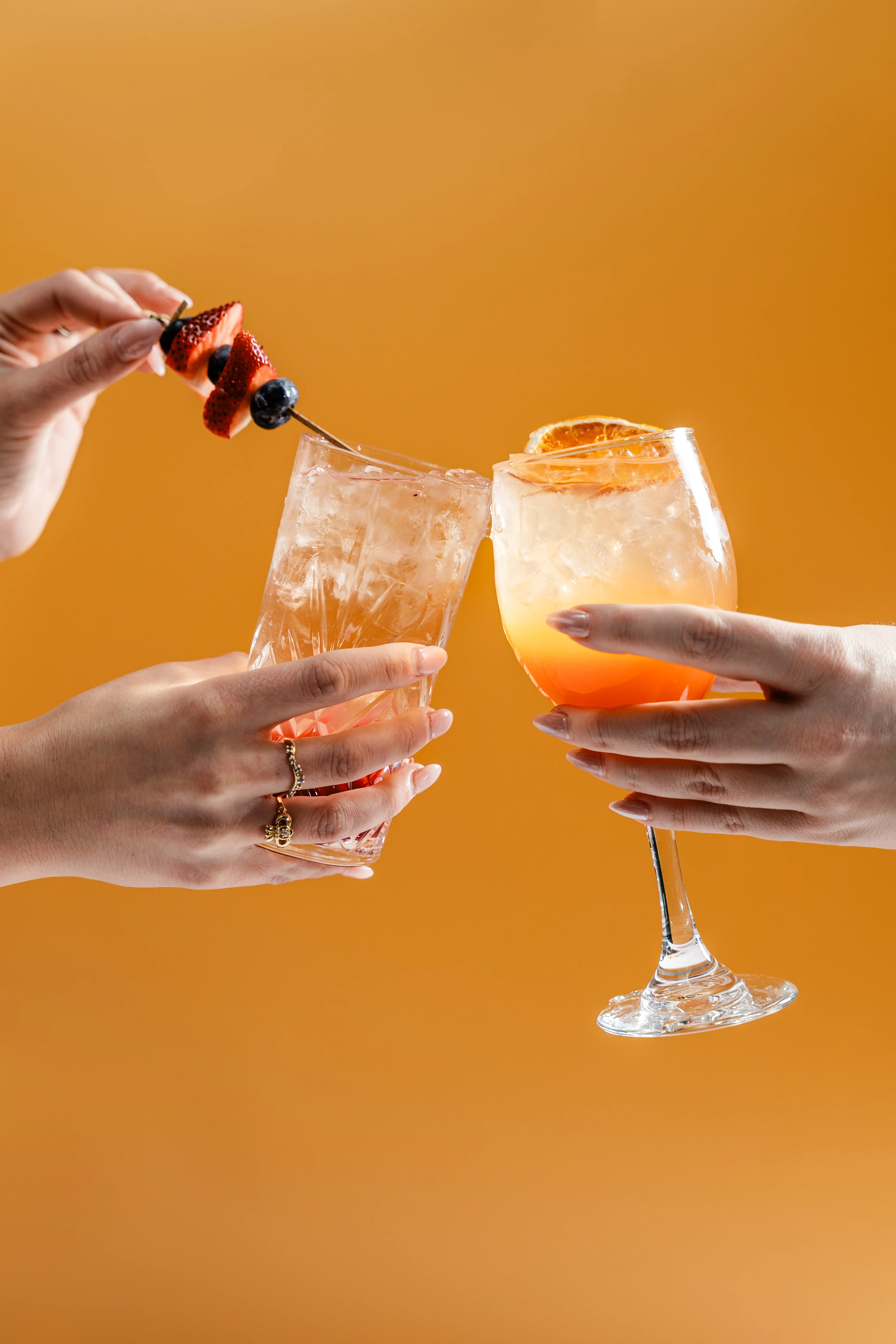 Two colorful cocktails clinking together with fruit garnishes against an orange backdrop