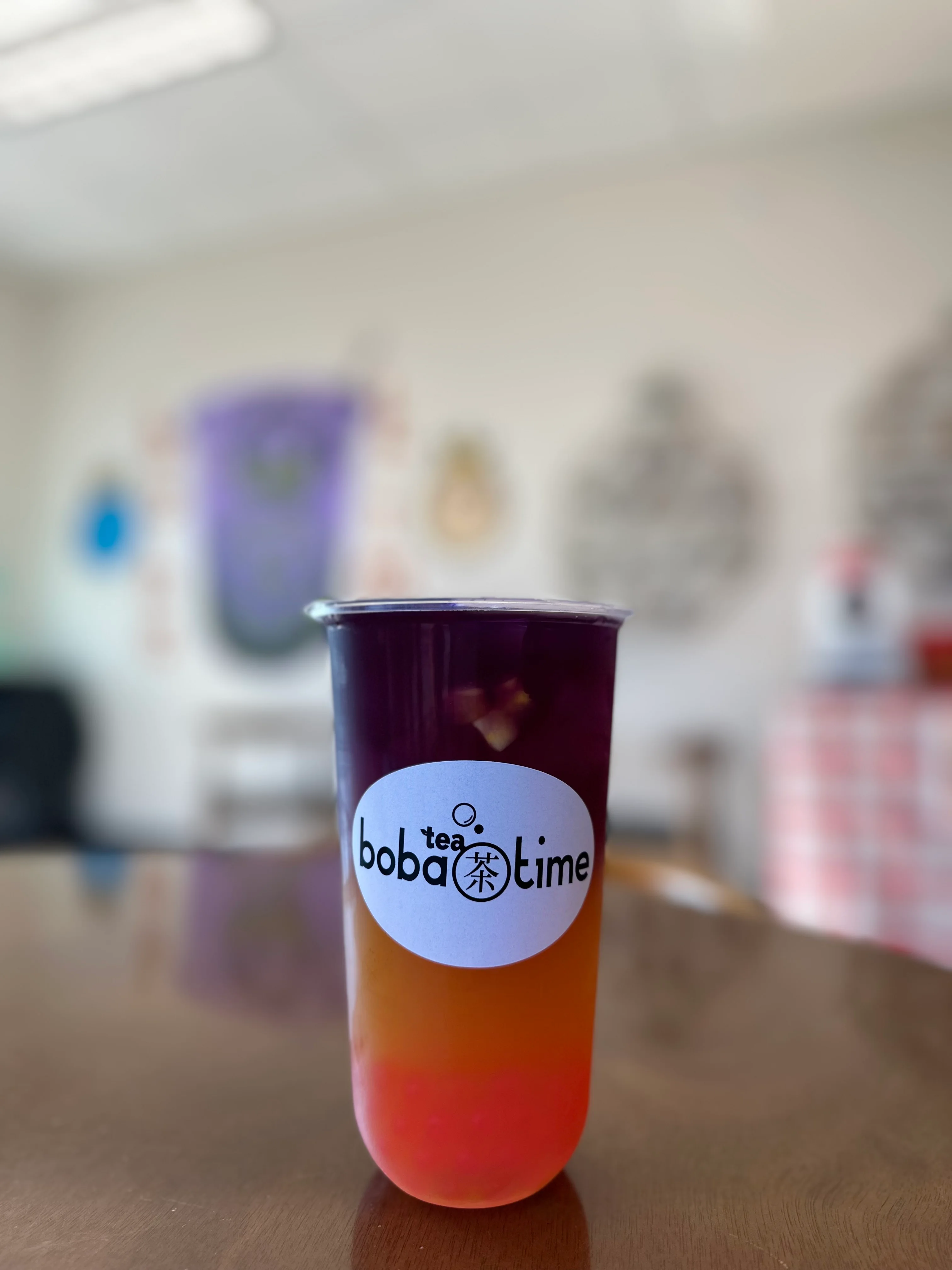A colorful layered drink served in a clear cup with a logo, featuring shades of purple and orange. The background includes a bright, modern café setting.
