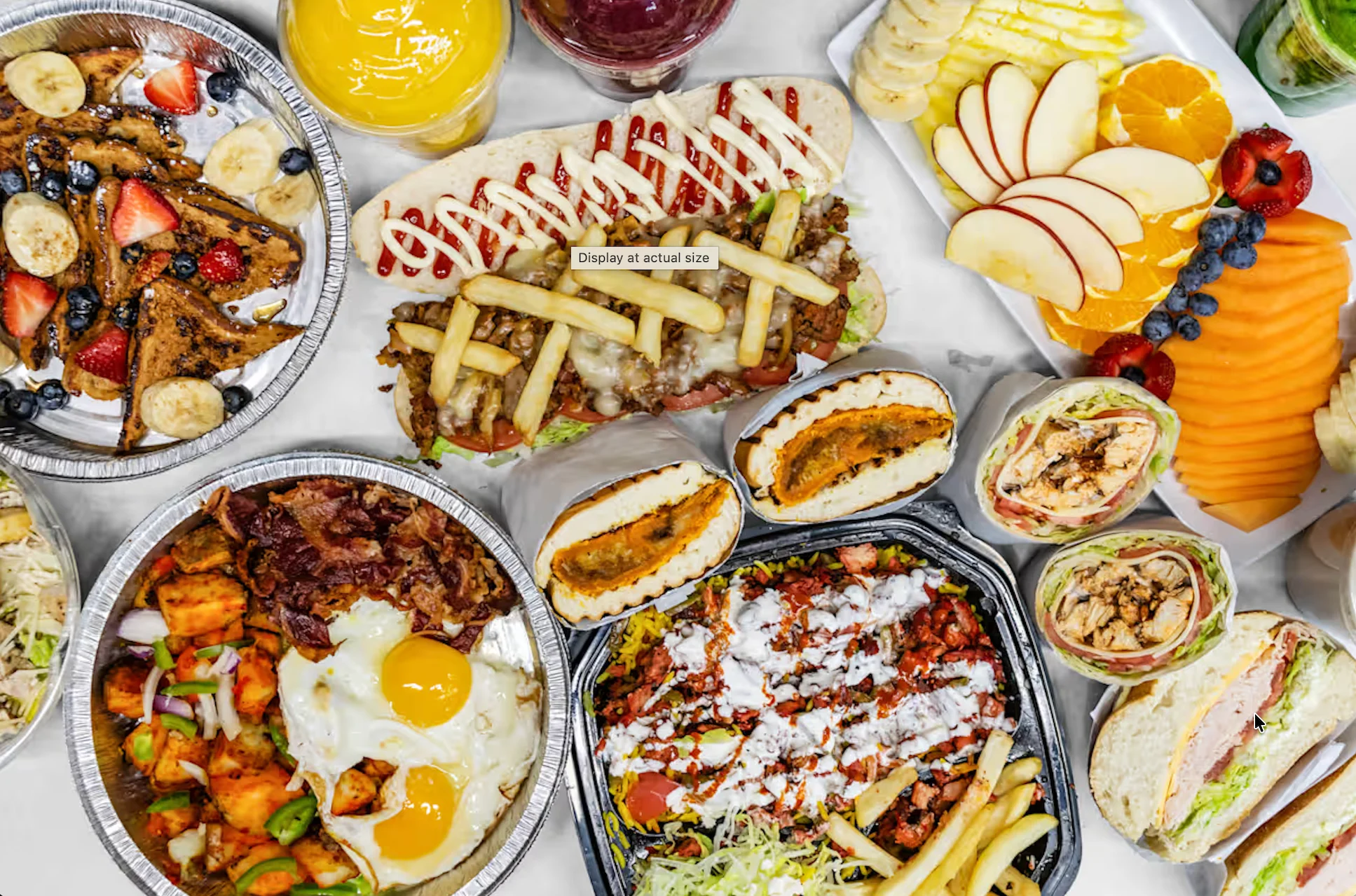 A vibrant spread of various dishes, including breakfast items like pancakes and eggs, sandwiches, wraps, and a colorful fruit platter featuring slices of apples, oranges, and berries.