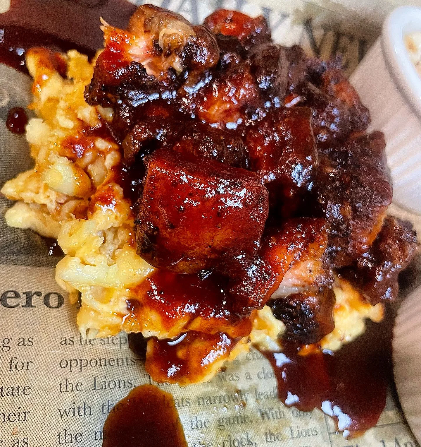 A serving of macaroni and cheese topped with pieces of smoked meat drizzled in barbecue sauce, displayed on newspaper.
