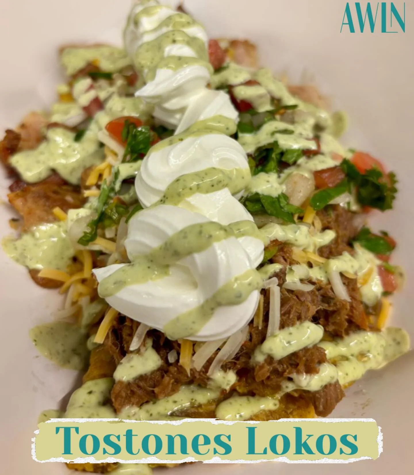 A plate of tostones topped with shredded meat, cheese, fresh vegetables, sour cream, and a creamy green drizzle.