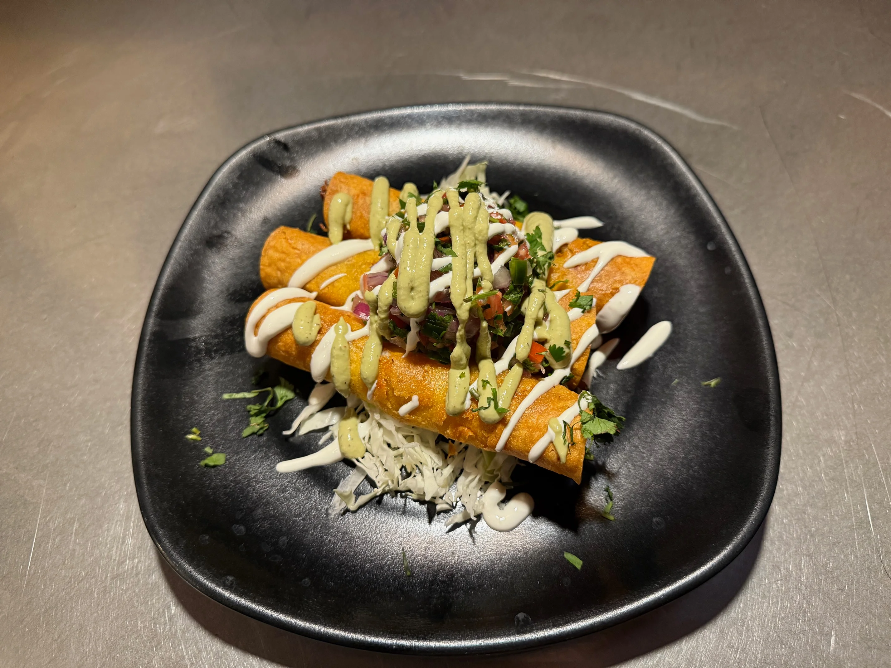 Three crispy taquitos topped with a drizzle of cream and avocado sauce, served on a bed of shredded lettuce.