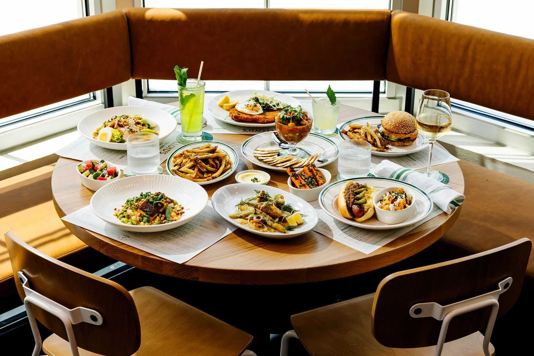 A spread of plated entrees, sandwiches, and appetizers on a light wood table at Lloyd's