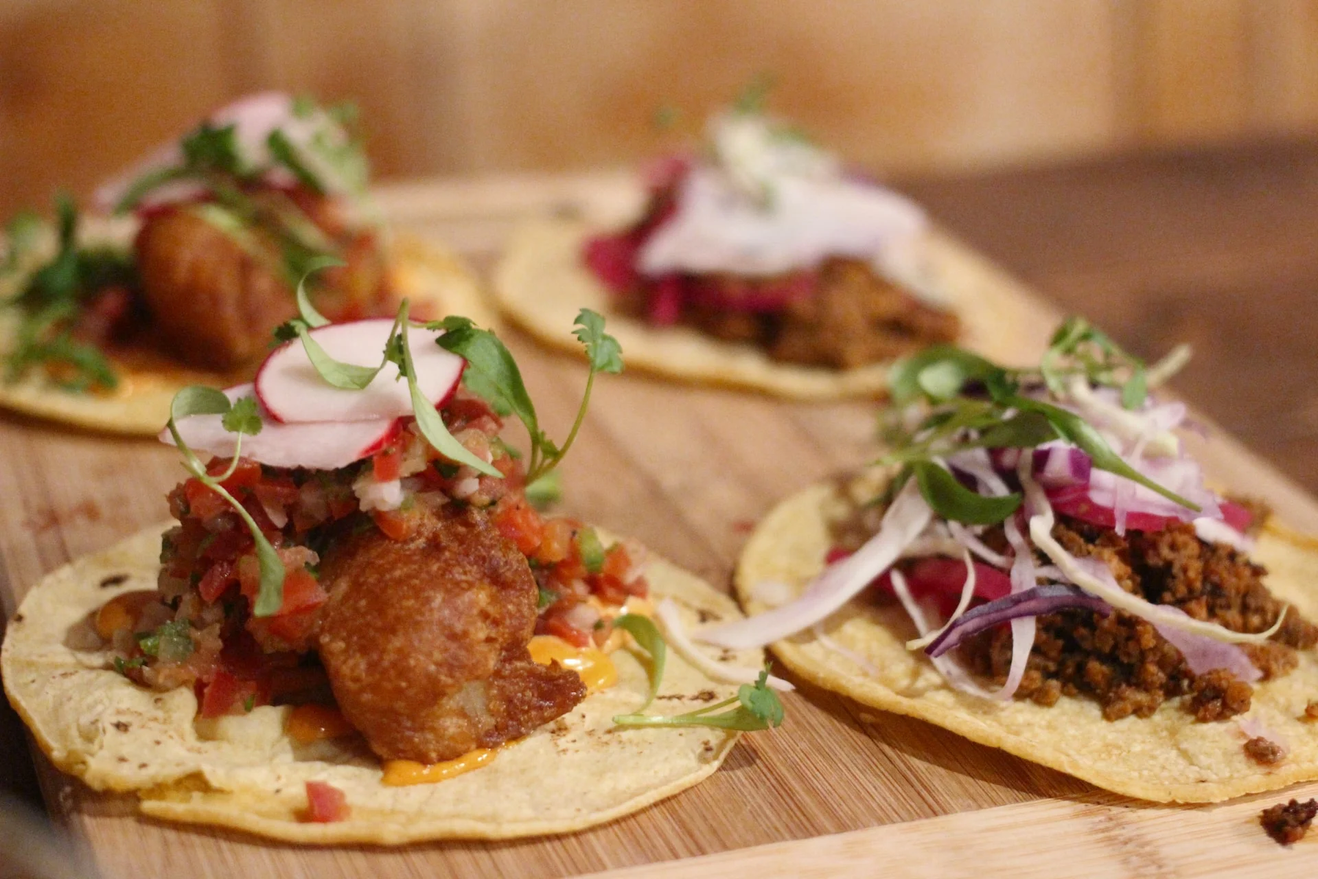 A selection of tacos arranged on a wooden board. Each taco is filled with ingredients such as sautéed meat, salsa, radishes, and fresh herbs, showcasing a vibrant display of colors and textures.