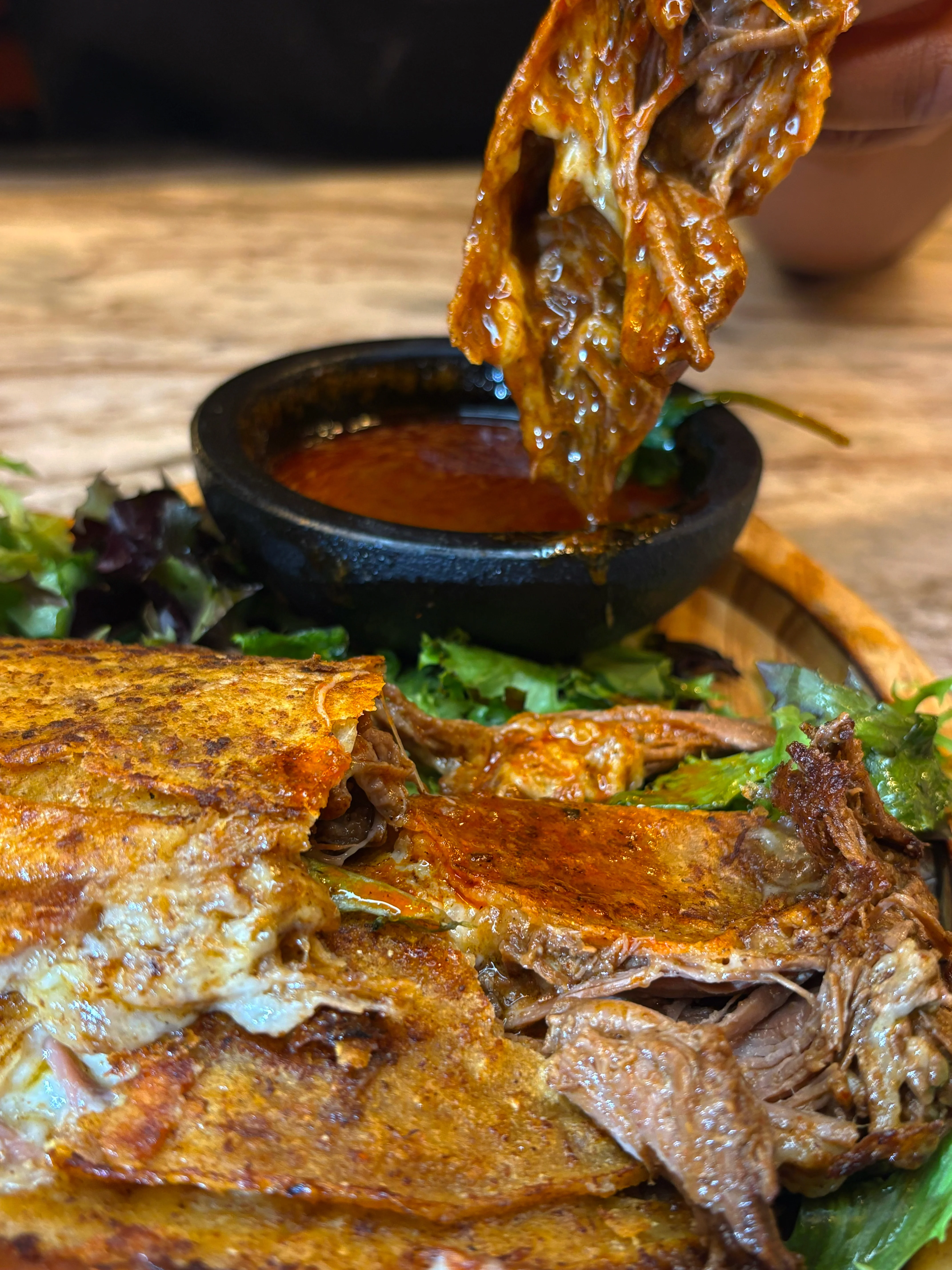 A close-up of a savory meat dish being dipped into a rich sauce, with a side of greens and flatbread.