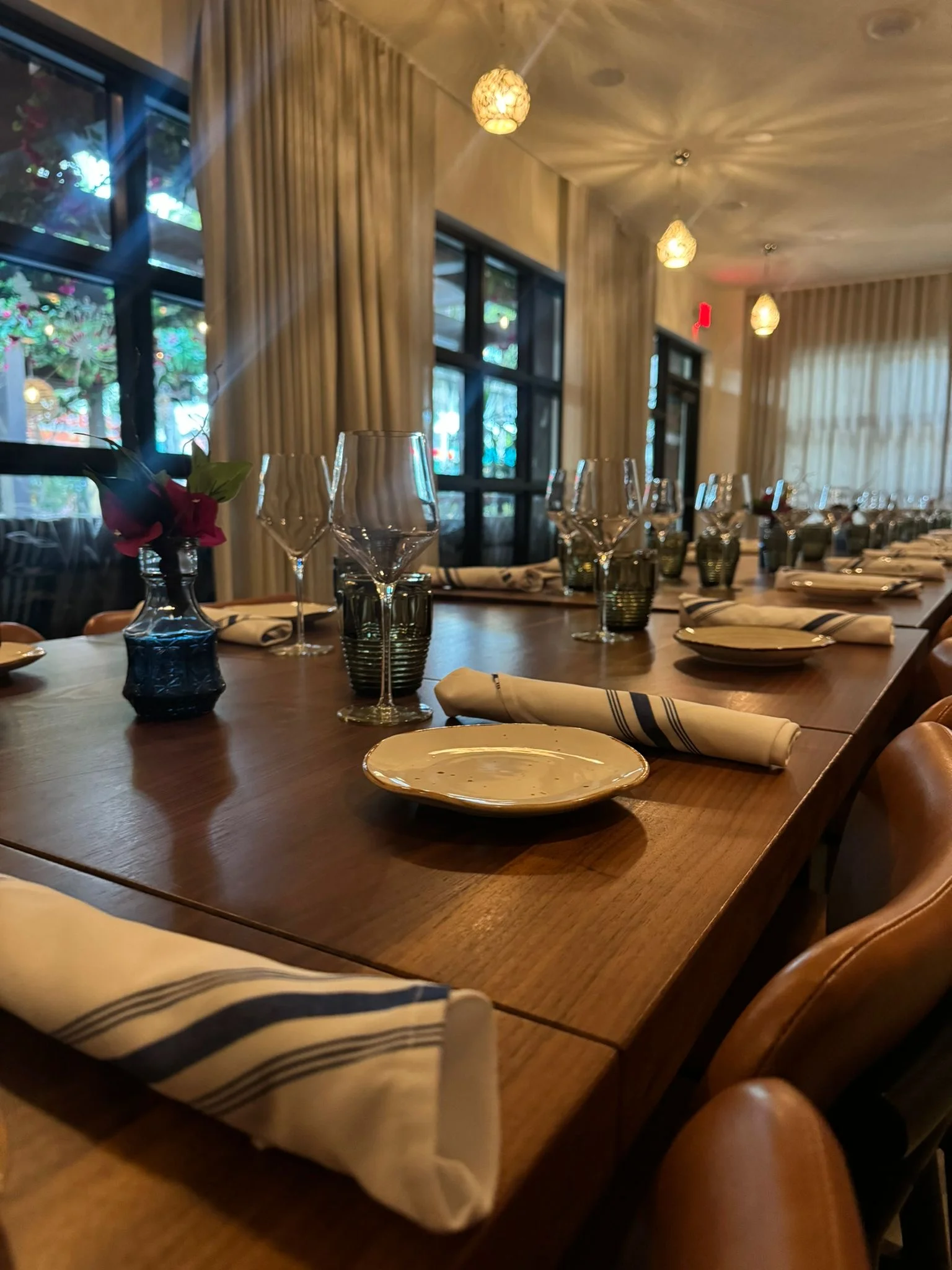 Close-up shot of a long wooden inning table with dining plate sets, juice and wine glasses with napkins neatly arranged on a long brown wooden table with rows of brown wooden dining chairs on both the right and left side of it.