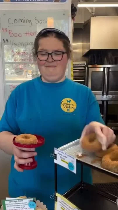 A bakery employee smiling while serving donuts. The counter is filled with various baked treats.