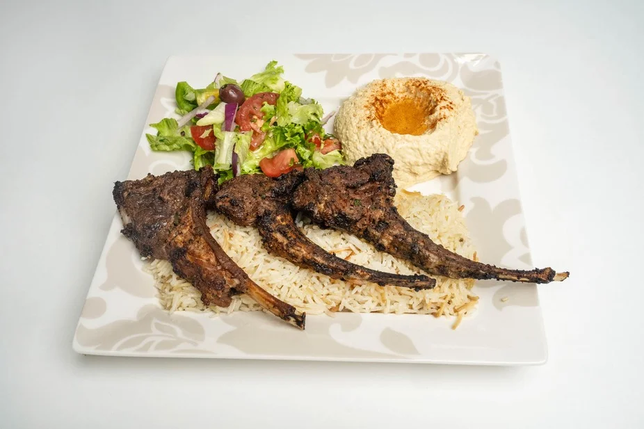 A plated meal featuring three grilled lamb chops served over rice, accompanied by a fresh salad and a portion of hummus. The dish is garnished with spices.
