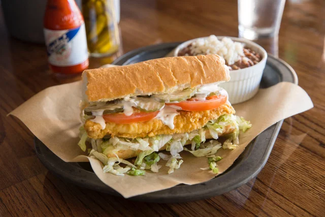 A sandwich filled with crispy fried shrimp, lettuce, and tomatoes, served with a side of stew in a bowl on a tray.