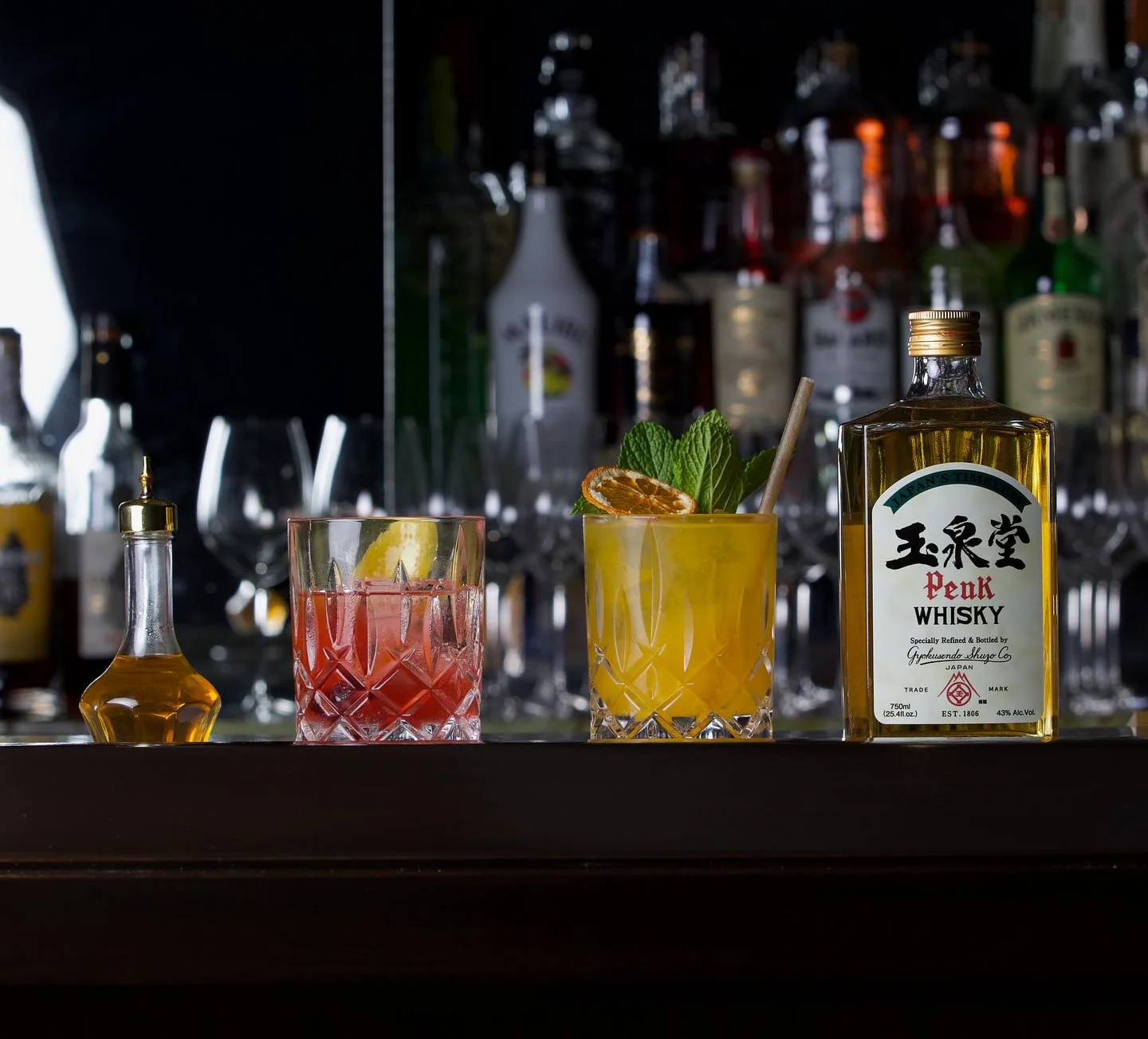 A bar counter display featuring a bottle of Peak Japanese Whisky, a yellow cocktail garnished with fresh mint and a dried citrus wheel, a red cocktail with a lemon twist in a crystal rocks glass, and a small glass bitter bottle, set against a blurred background of various liquor bottles.