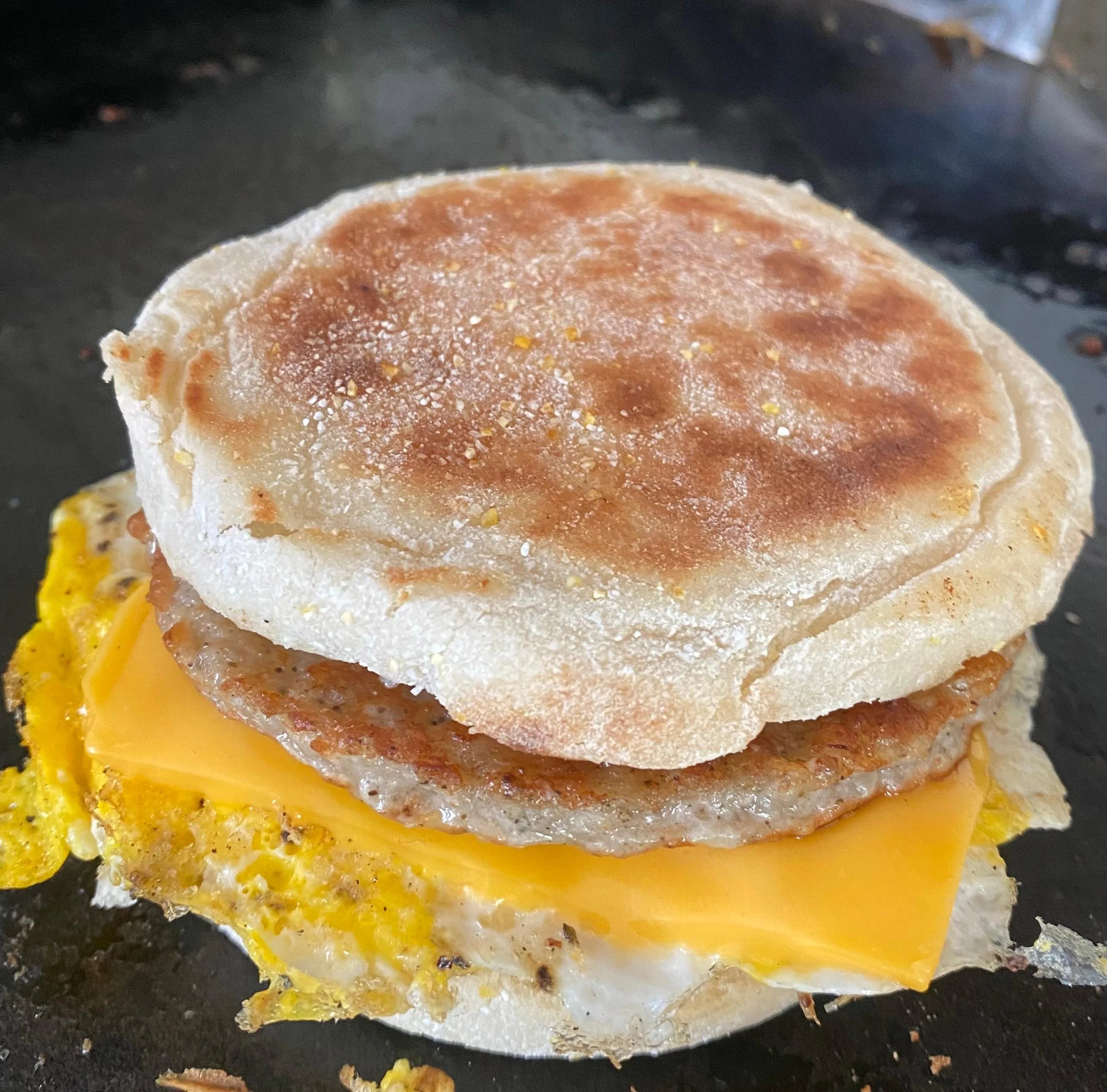 A toasted English muffin sandwich with cheese, egg, and sausage, captured on a griddle.