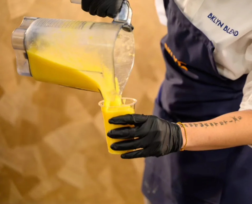 Person in black gloves pours yellow smoothie from a blender into a clear cup. They're wearing a dark apron. The scene feels professional and focused.