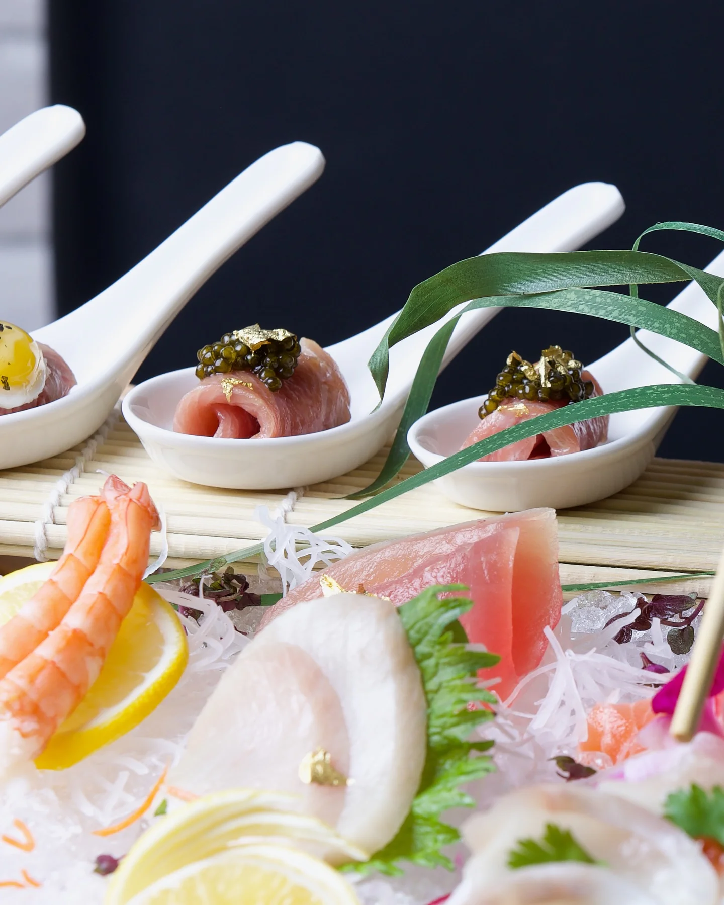 A close-up of an upscale sashimi and appetizer presentation, featuring small rolls of raw fish topped with caviar and gold leaf in white ceramic tasting spoons, arranged above an assortment of fresh sashimi, including shrimp, tuna, and white fish, beautifully served over shaved ice with lemon slices and daikon radish strands.