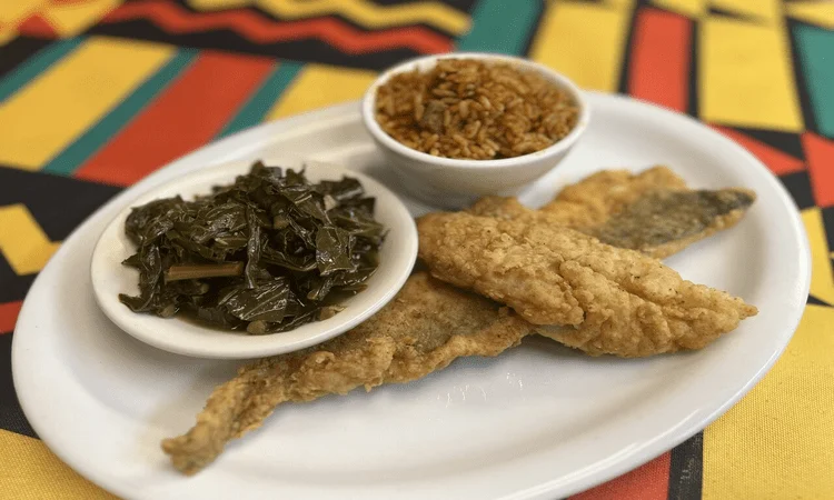 A dish consisting of fried fish, a serving of seasoned rice, and a side of cooked greens, set against a vibrant tablecloth.