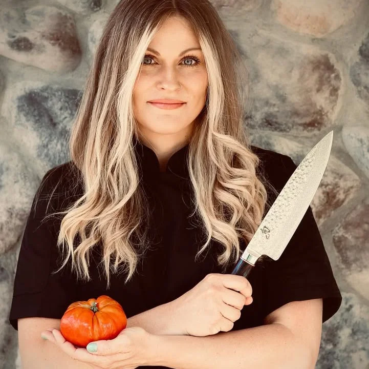 A person holding a tomato in one hand and a large kitchen knife in the other, standing against a stone wall.