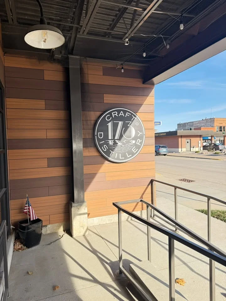 The exterior of a modern craft distillery, featuring a large logo on a wooden panel, with visible signage and an American flag nearby.