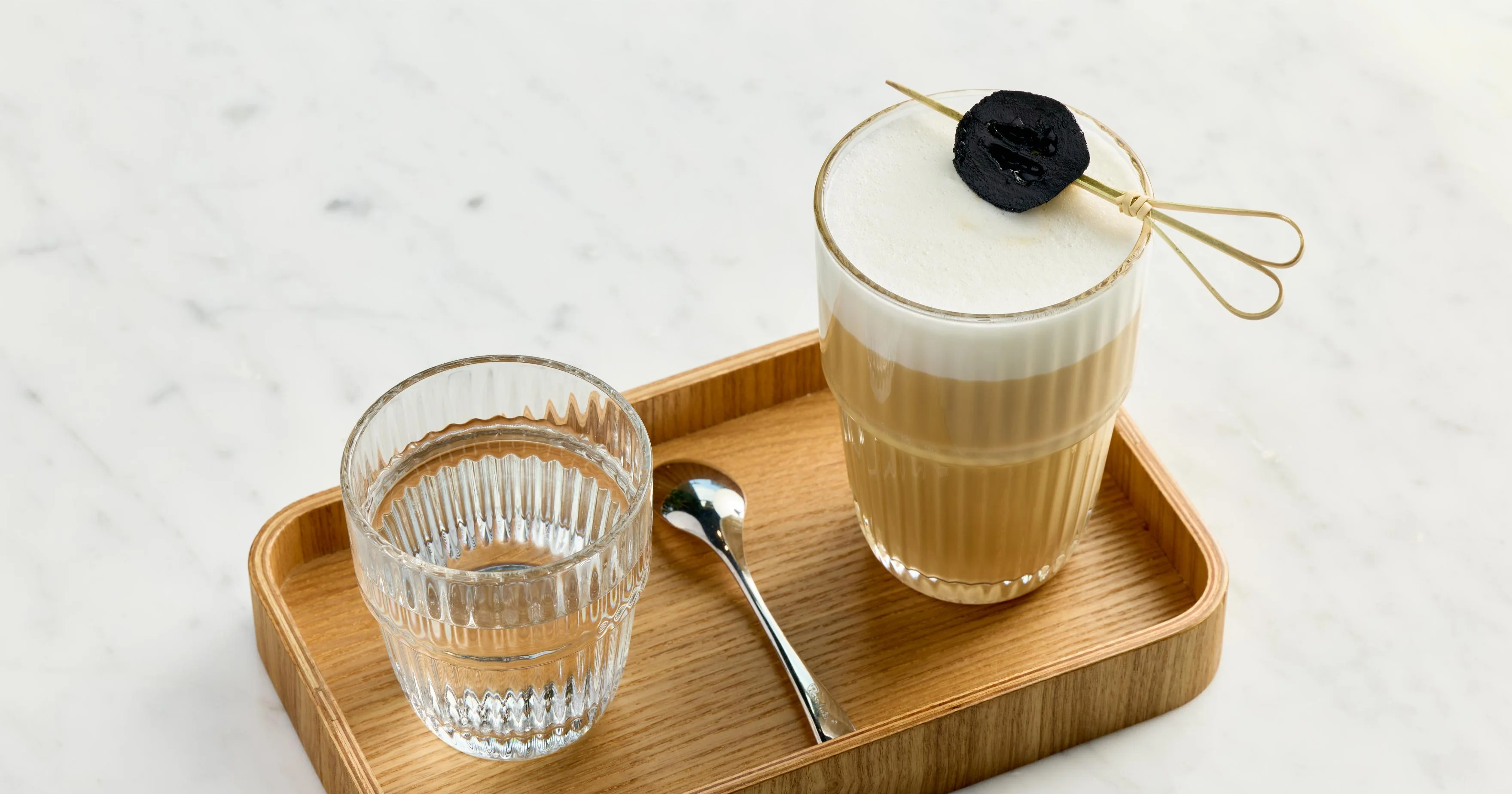 A glass of coffee with foam and a glass of water on a wooden tray.