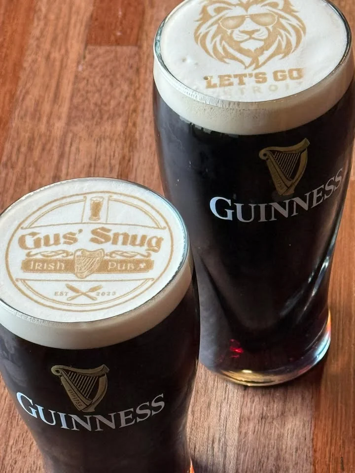 Two pints Guinness, featuring detailed foam art on top, set on a wooden table.