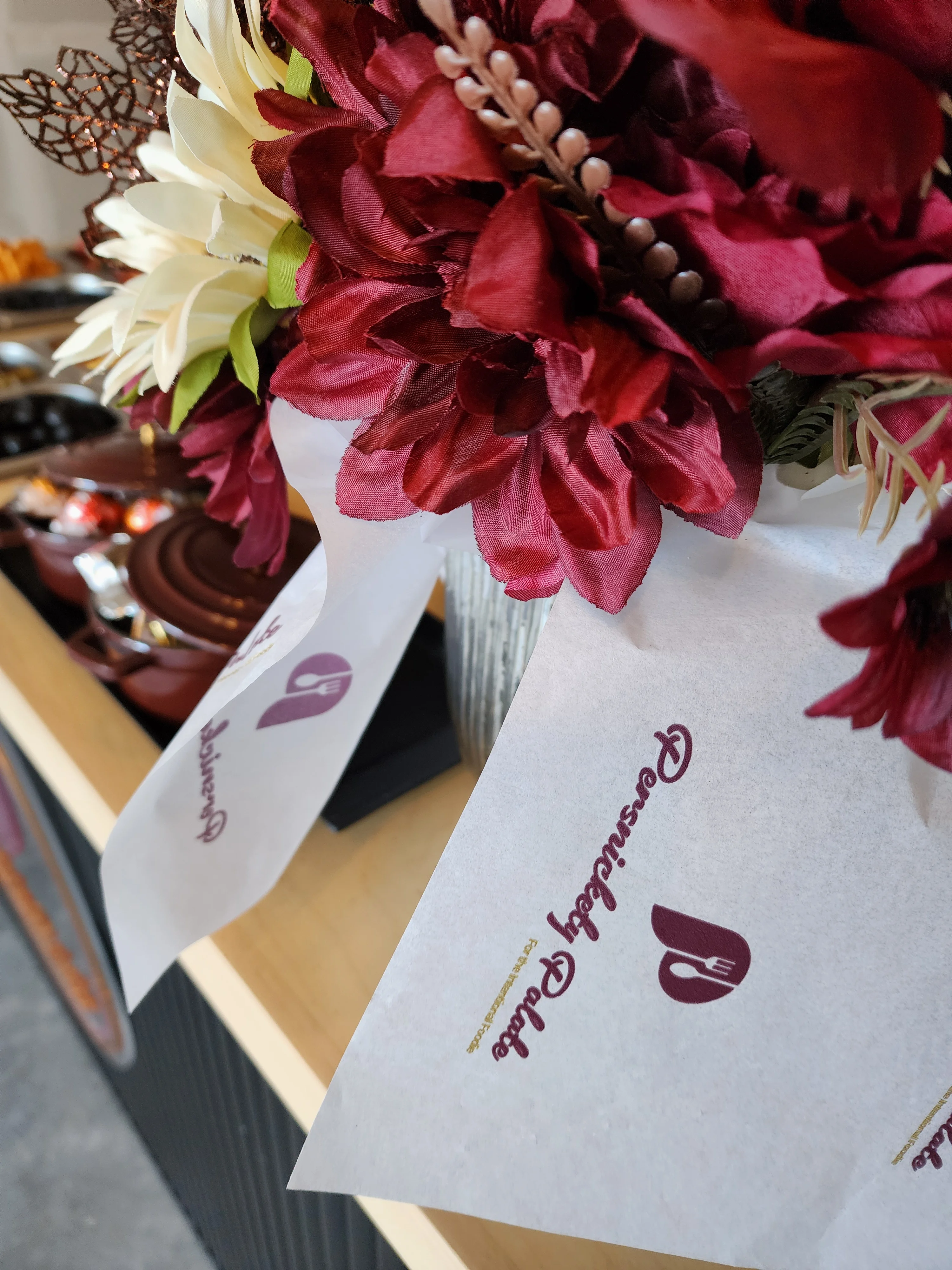 A decorative arrangement of artificial flowers in shades of burgundy and cream, with elegant ribbons attached. The background features a buffet table with food items.