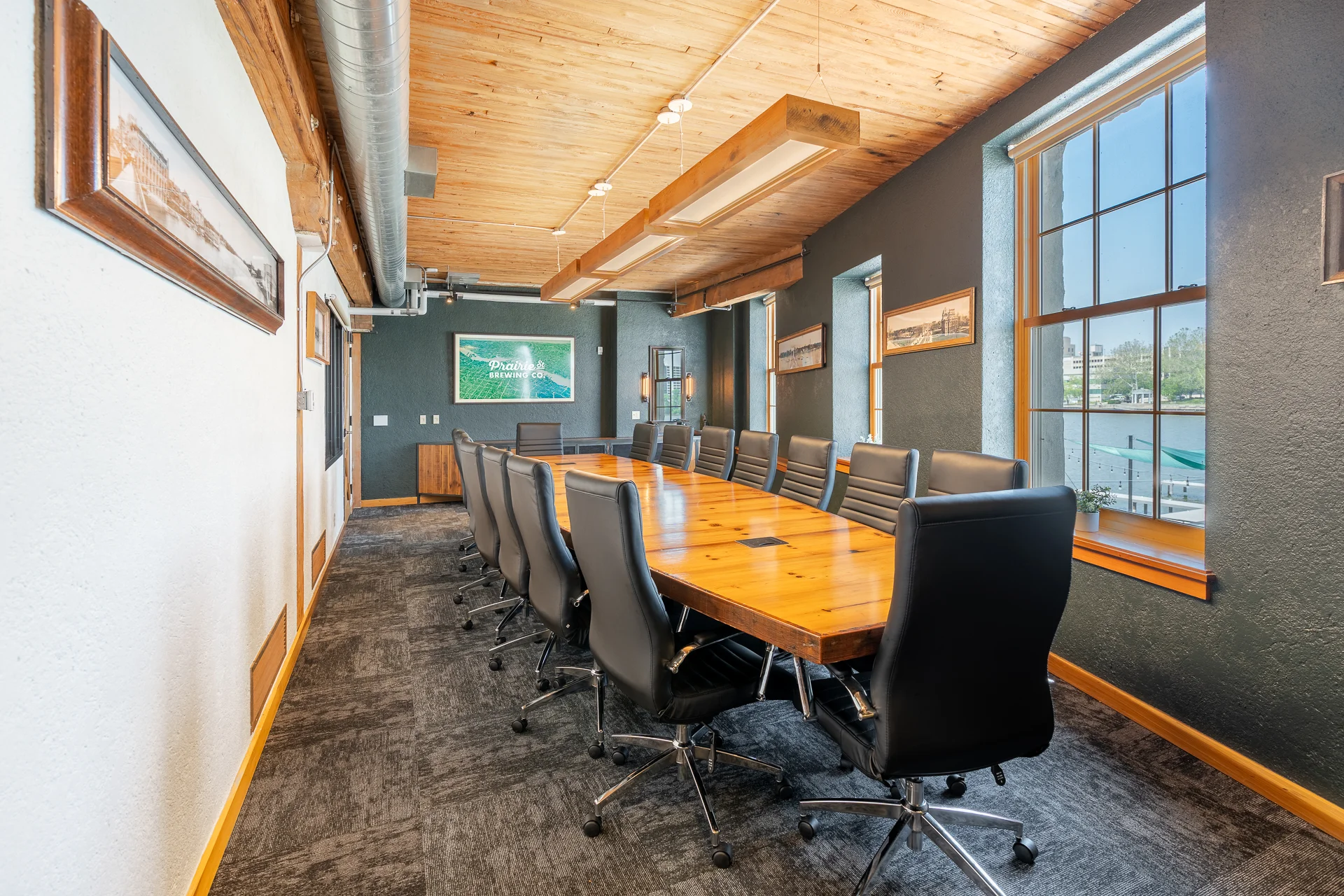 A modern conference room with a long wooden table surrounded by black leather chairs, illuminated by natural light from large windows.