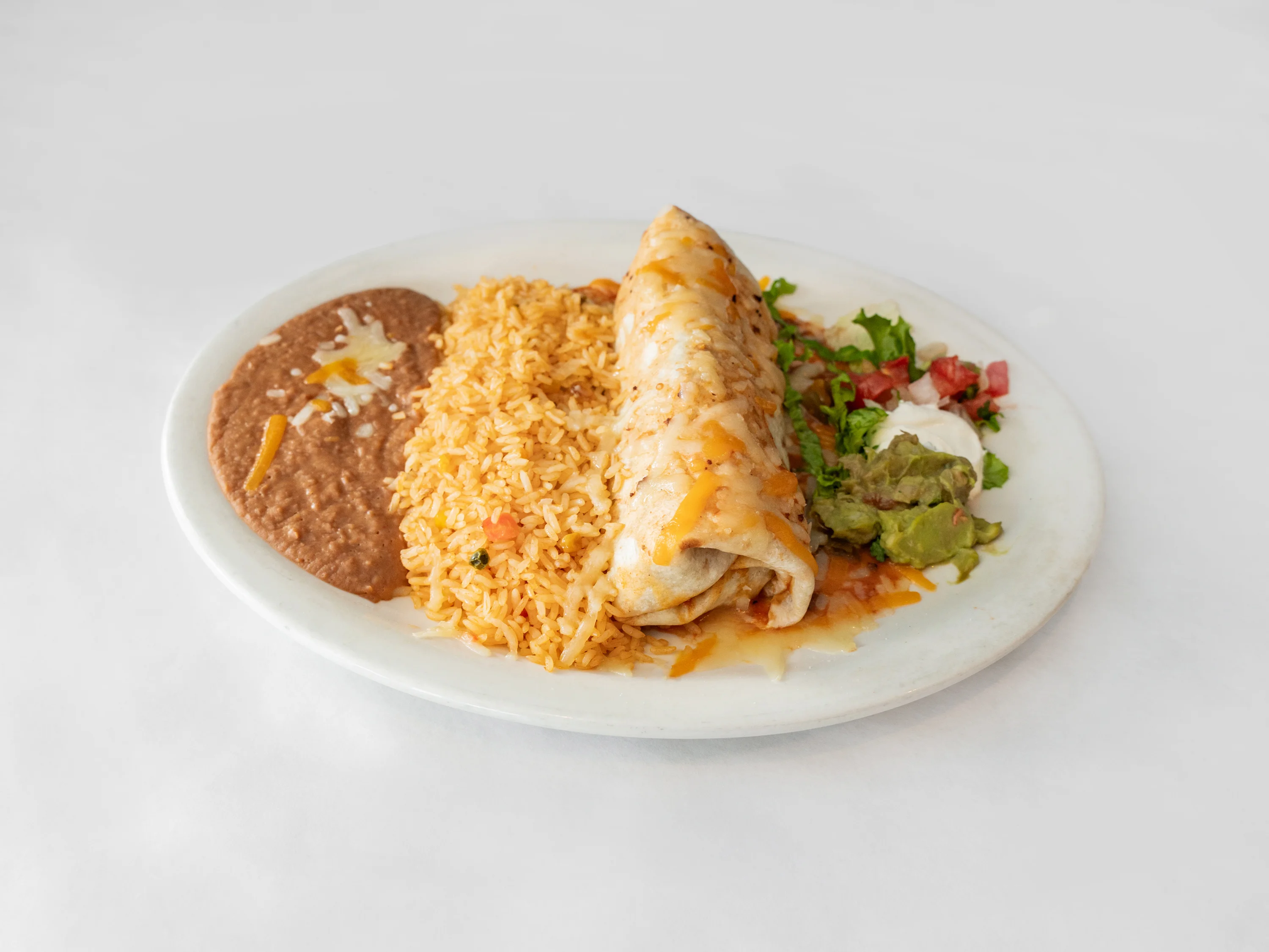 A plate of a burrito with rice and refried beans, accompanied by guacamole, lettuce, tomato, and sour cream.