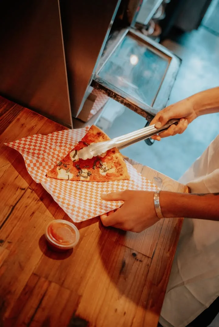 A slice of pizza is being served with tongs on a checkered paper. A small cup of sauce is placed beside it.