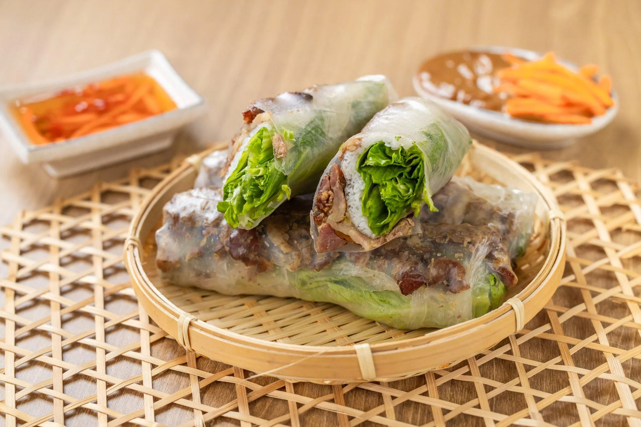 A bamboo basket filled with rolled rice paper wraps containing lettuce and meat. Accompanying sauces and pickled vegetables are visible in the background.