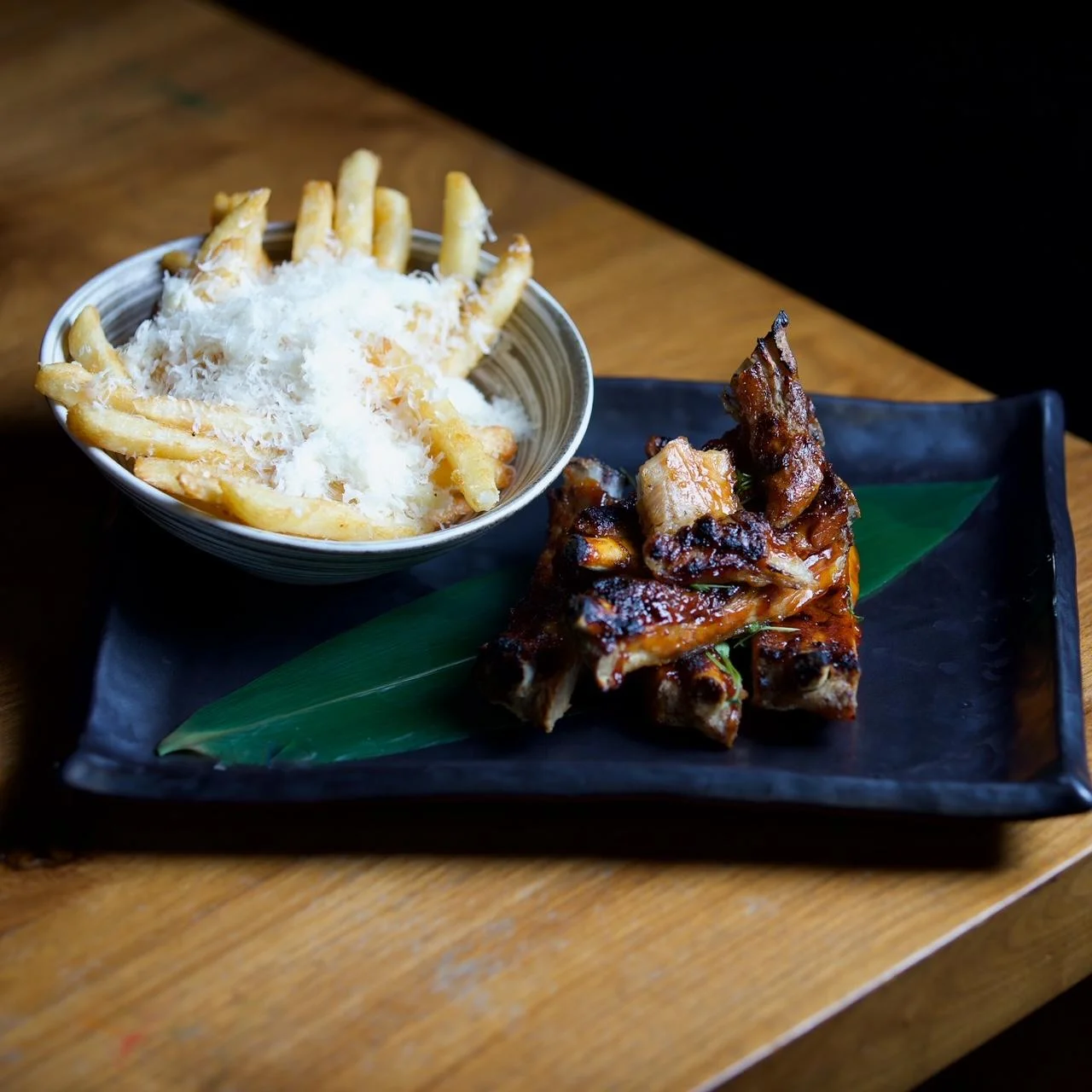 A small stack of glazed, grilled ribs arranged on a green leaf atop a dark square plate, served alongside a bowl of golden french fries heavily dusted with grated parmesan cheese on a wooden table.