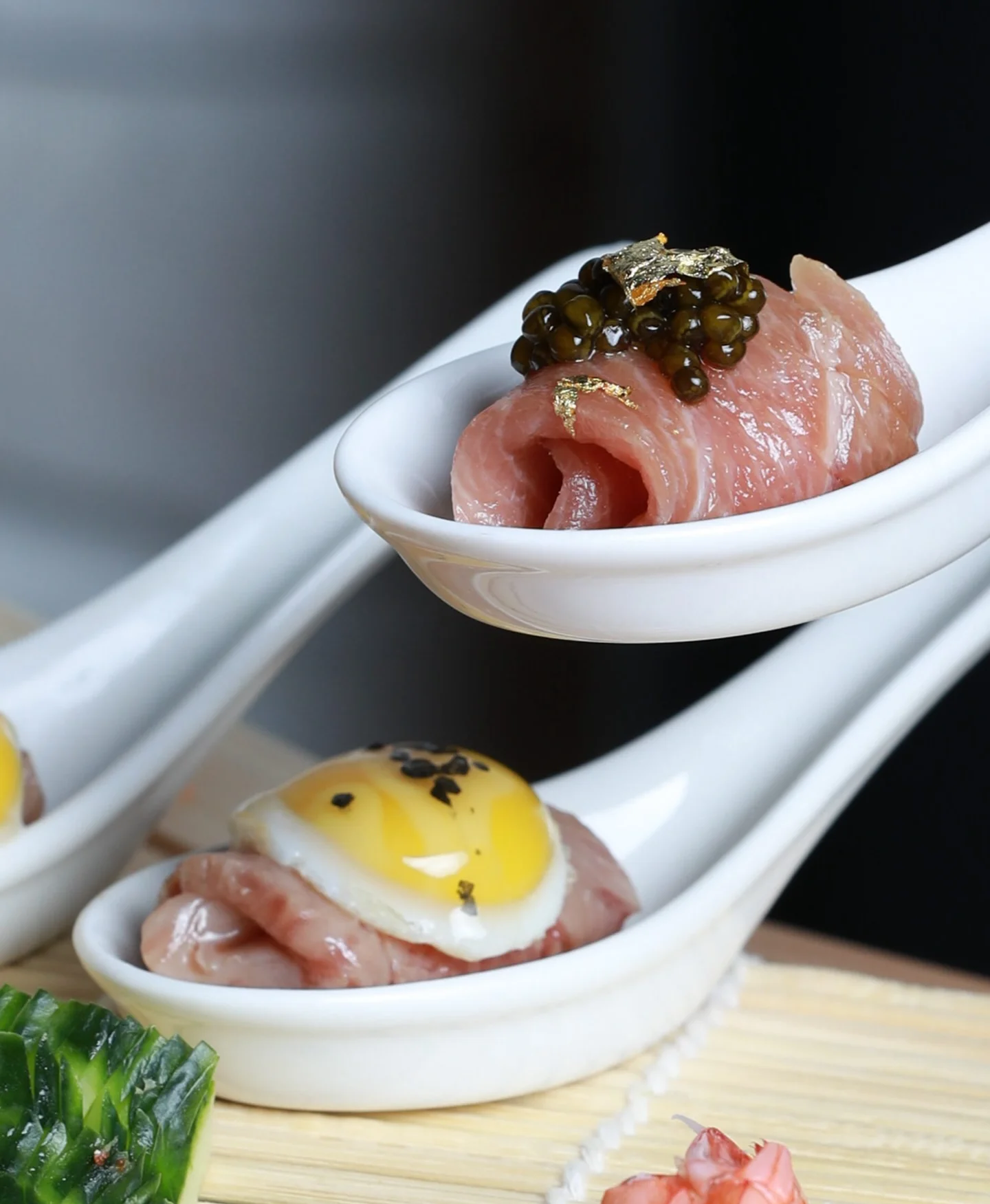 A close-up of gourmet appetizers presented in white ceramic tasting spoons, showcasing a rolled piece of raw fish topped with black caviar and gold leaf in the foreground spoon, and another piece of meat topped with a miniature fried egg in the background spoon.
