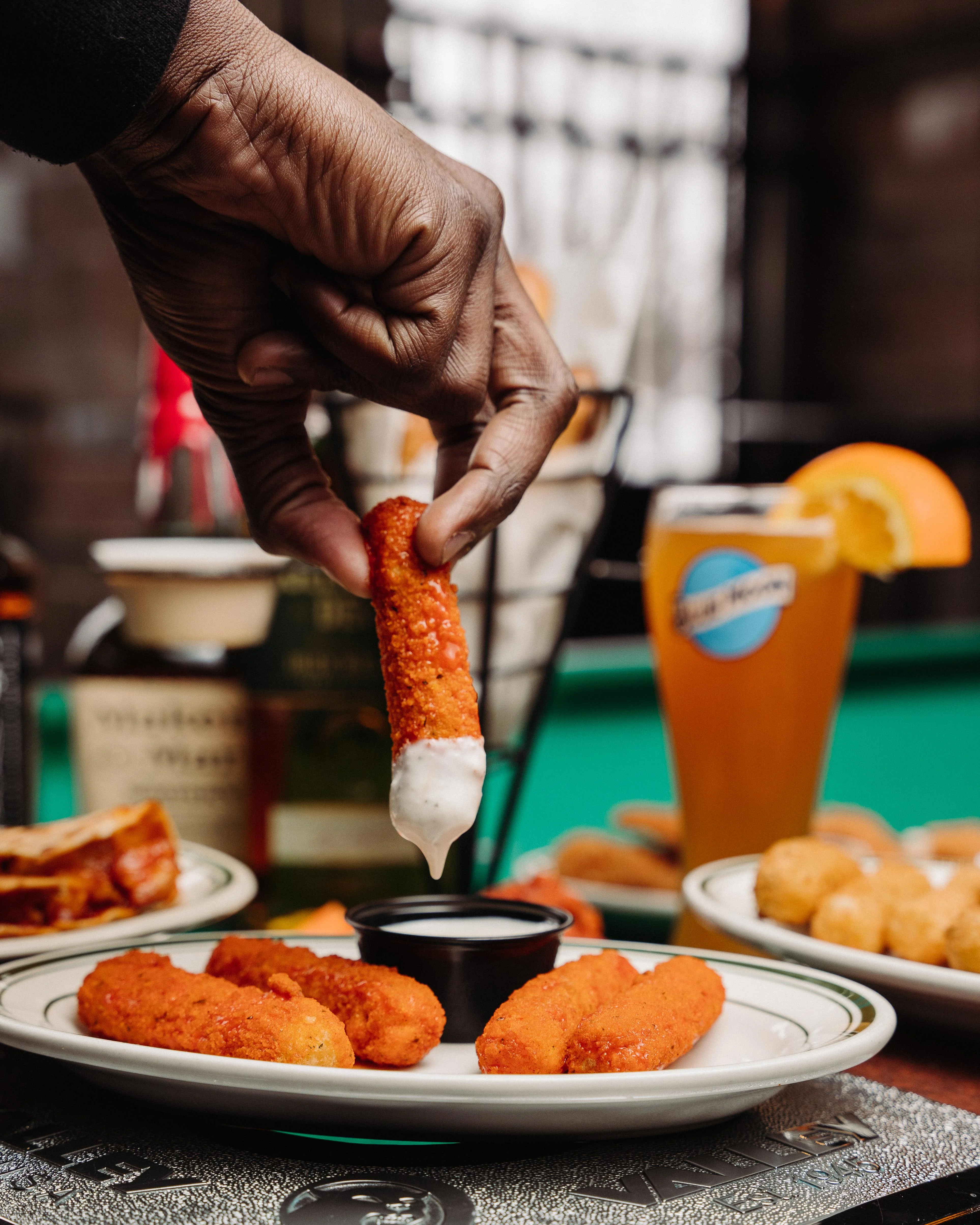 A hand dips a crispy orange snack stick into creamy white sauce. In the background, a drink and various appetizers are visible.