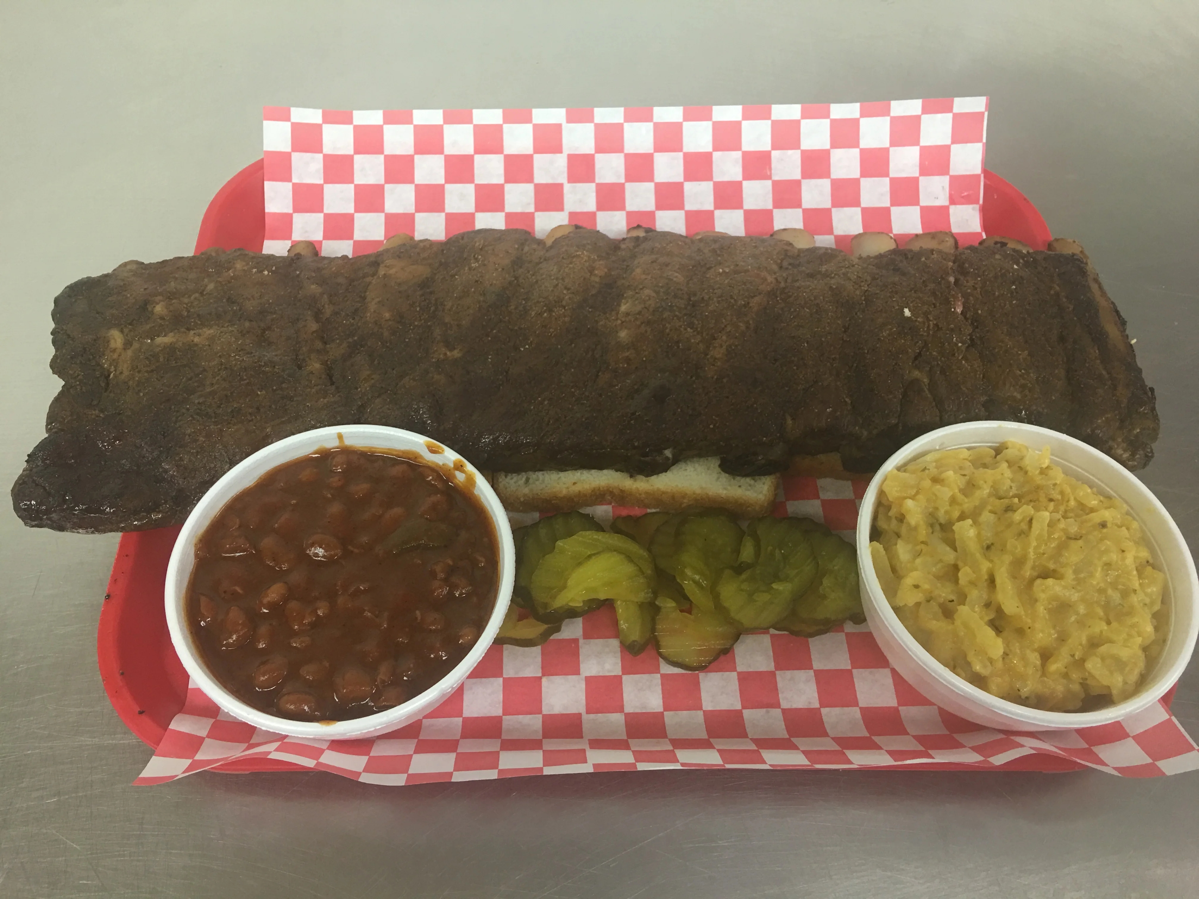 A barbecue platter featuring ribs, baked beans, pickles, and macaroni and cheese on a red checkered tray.