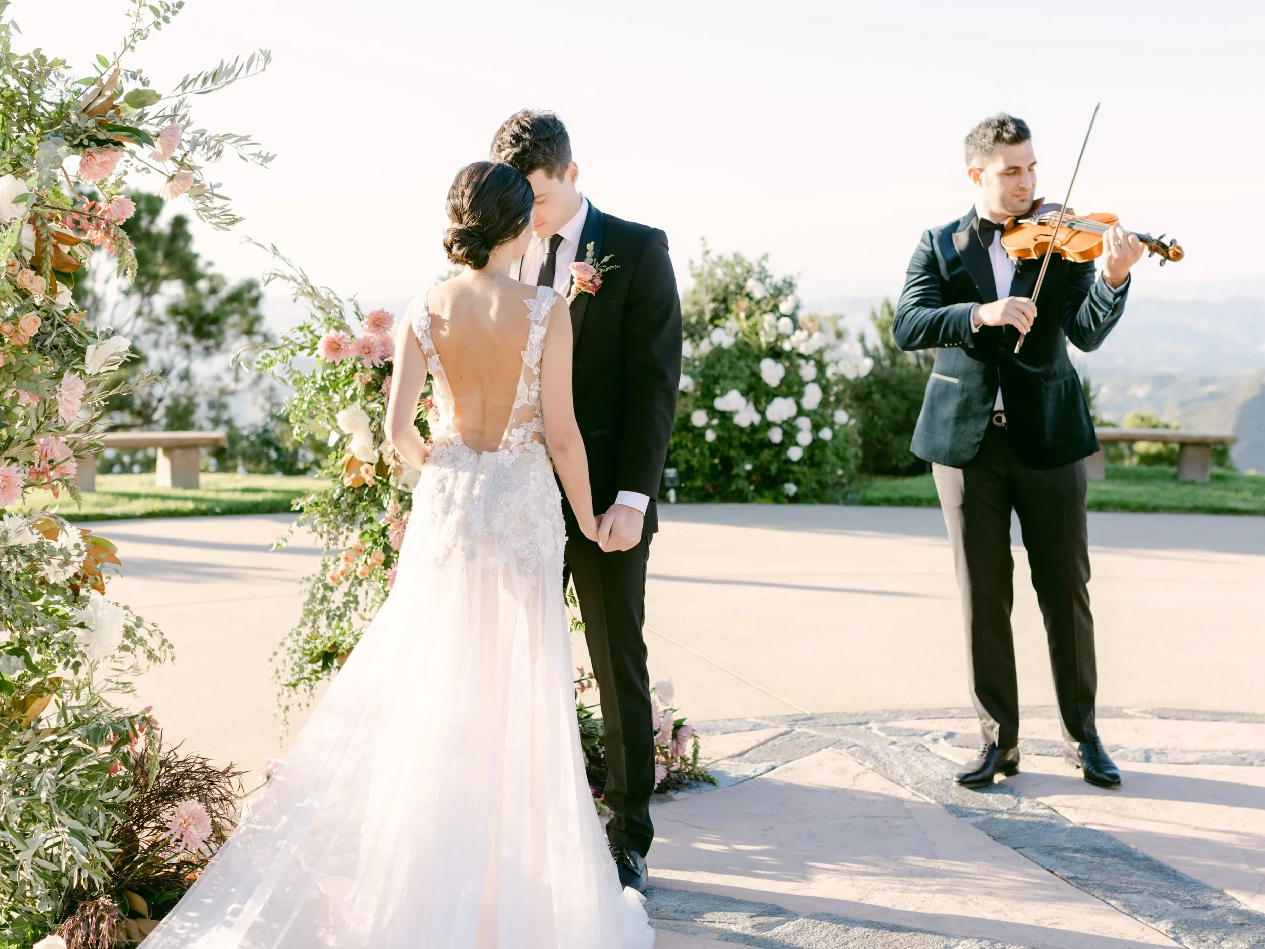 A wedding ceremony taking place outdoors, featuring a couple holding hands and a violinist playing nearby, surrounded by floral decorations.