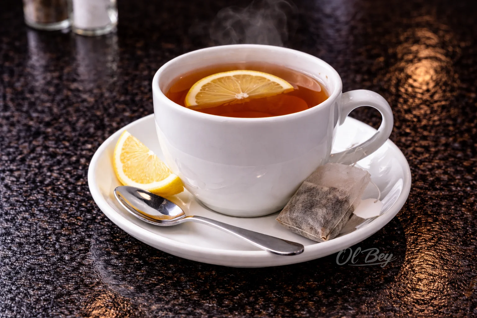 A steaming cup of tea garnished with a slice of lemon, served on a white plate alongside a tea bag and a silver spoon.