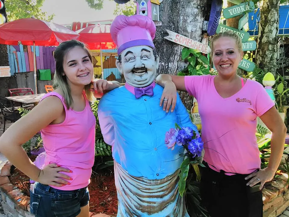 Two women posing with a colorful chef statue in a vibrant outdoor restaurant setting