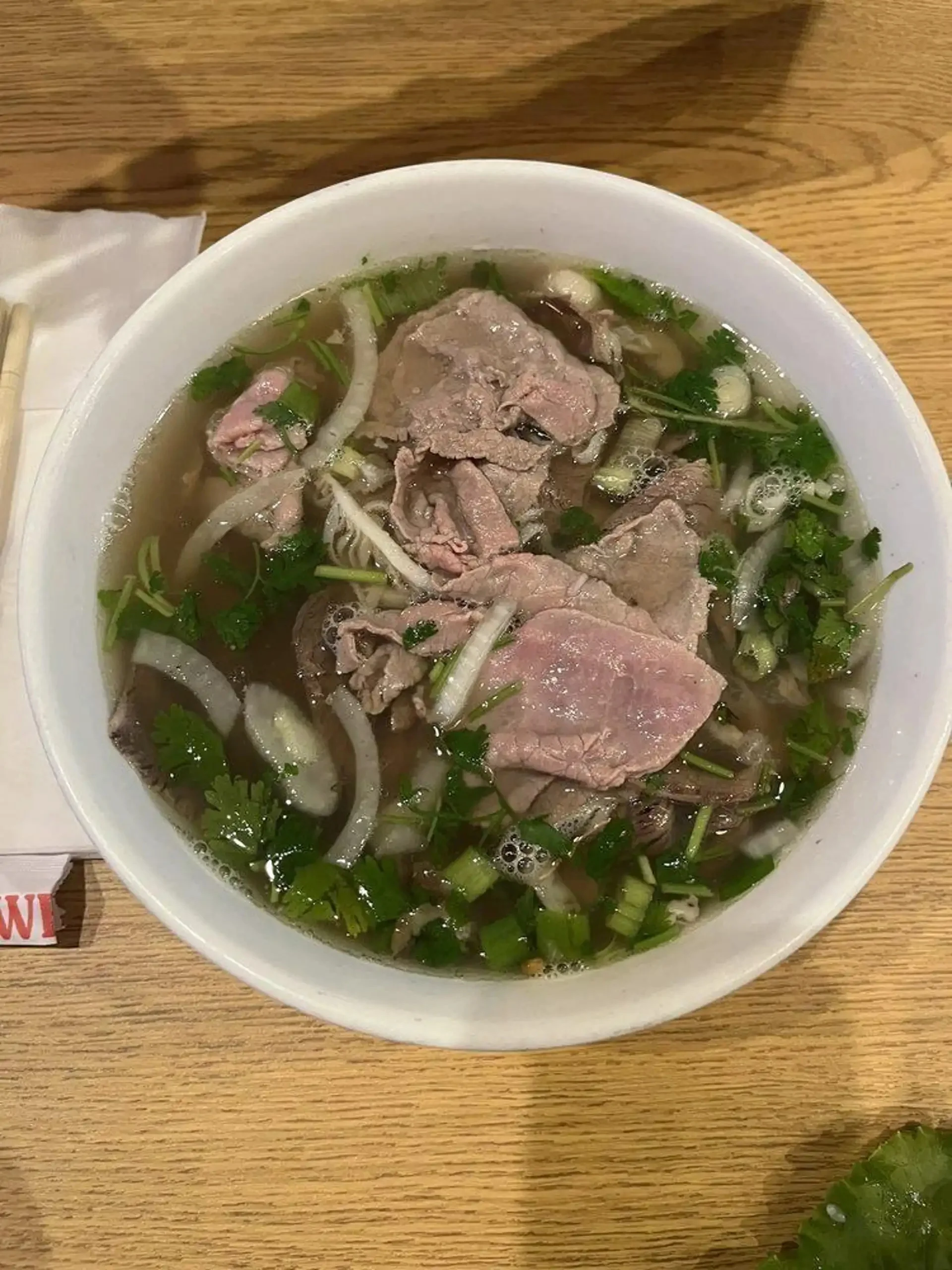 A bowl of pho featuring slices of beef, fresh herbs, and clear broth, garnished with cilantro and green onions.