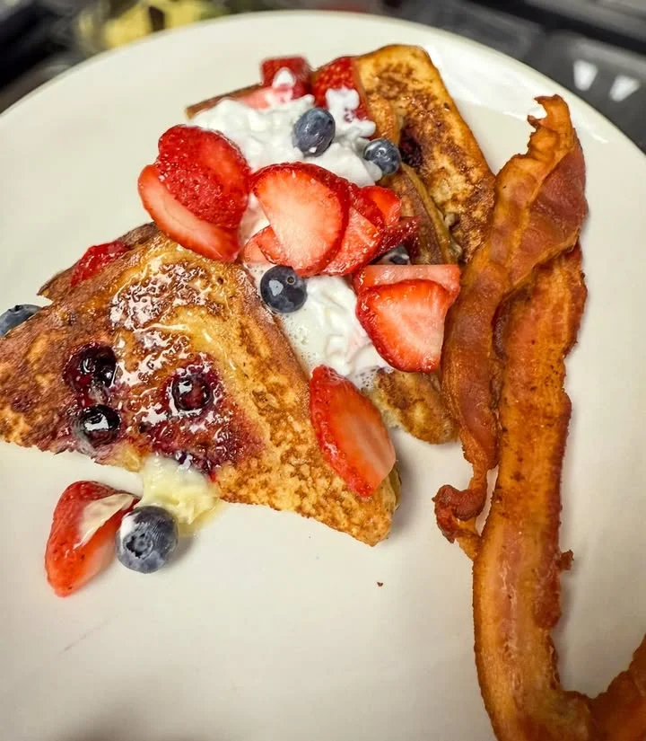 Delicious French toast topped with fresh strawberries, blueberries, and a dollop of whipped cream, accompanied by crispy bacon.