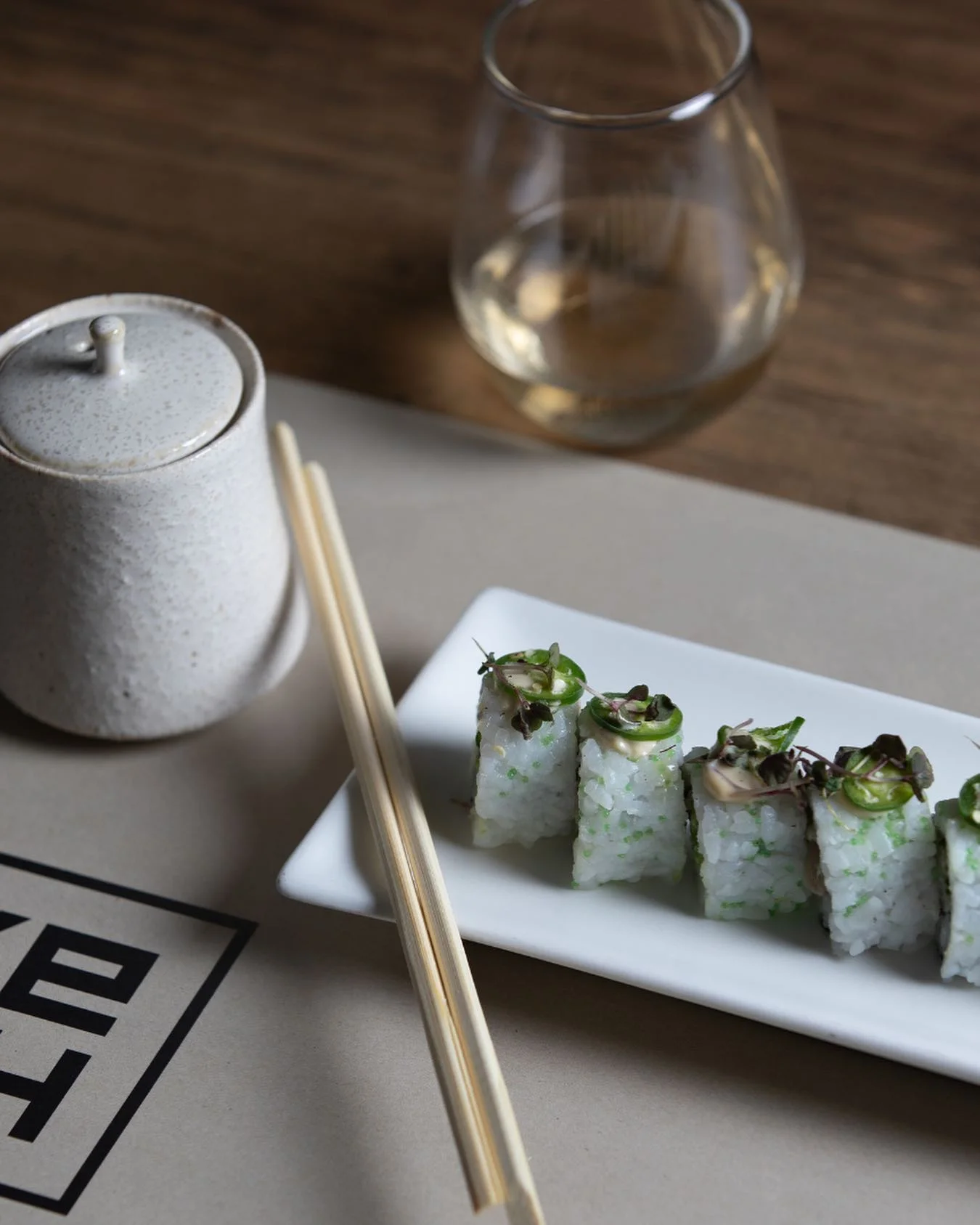 A row of sliced sushi rolls with green tobiko visible in the rice, topped with spicy mayo, jalapeño slices, and microgreens, served on a white rectangular plate alongside wooden chopsticks, a speckled ceramic soy sauce container, and a glass of white wine on a restaurant table.