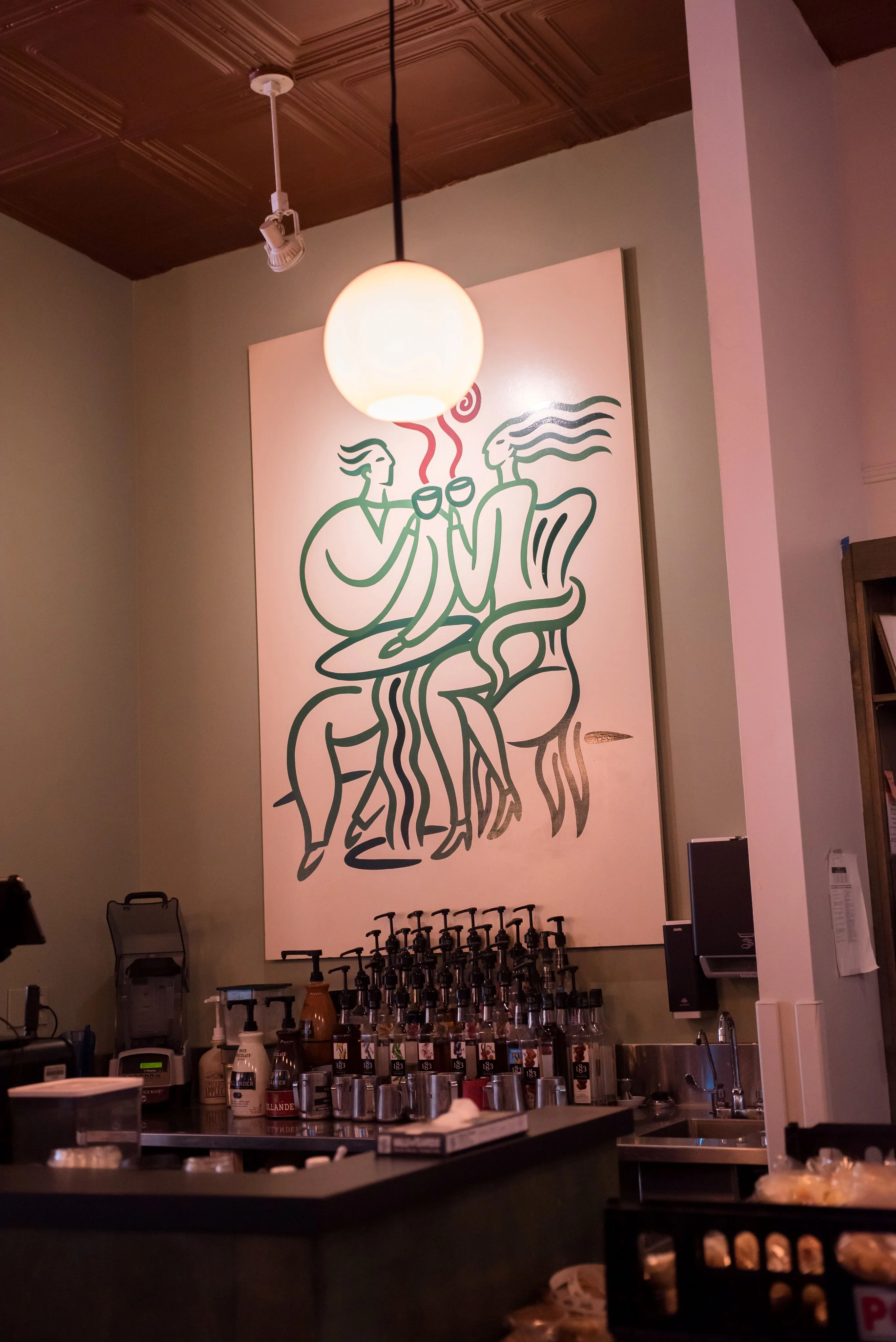 A café interior features a modern outline painting of two figures drinking together, set above a counter with syrup bottles and a warm light. Cozy and inviting.