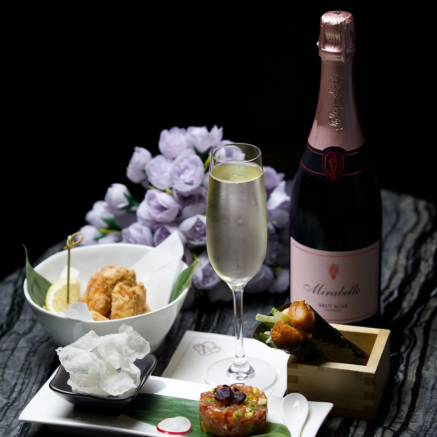 A premium dining spread featuring a bottle of Mirabelle Brut Rosé champagne next to a filled champagne flute, a crispy temaki hand roll in a wooden box, a small platter of tuna tartare with rice crackers, and a bowl of fried chicken karaage with a lemon wedge, set against purple flowers and a dark marbled surface.