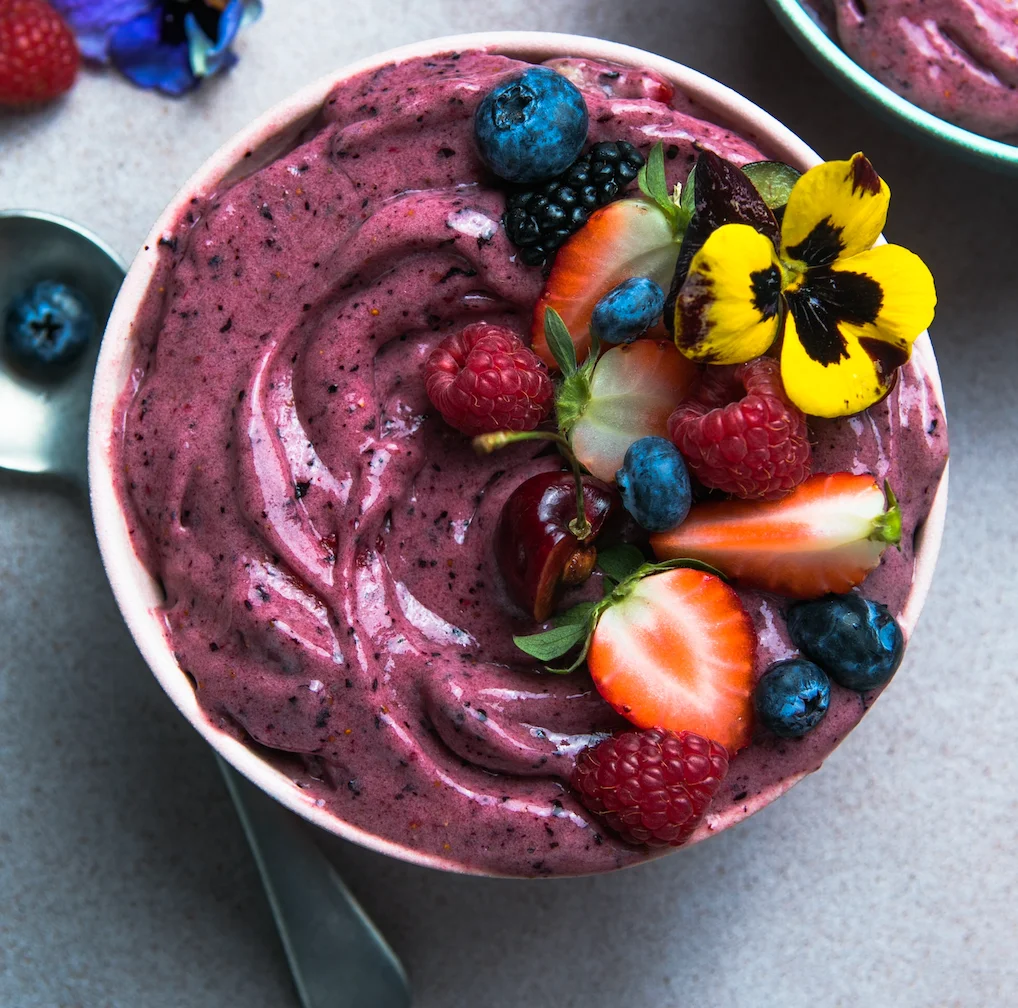 A creamy purple dessert bowl featuring a blend of berries, topped with strawberries, raspberries, and an edible flower.