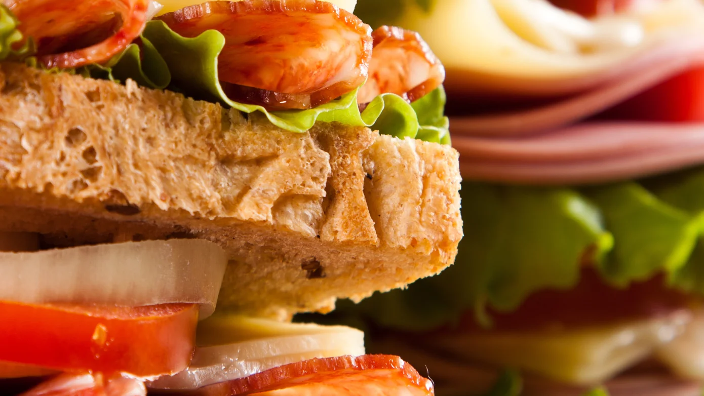 Stacked deli sandwiches with whole grain bread, sliced meats, cheese, lettuce, cucumbers, onions, and tomatoes, layered for a fresh and hearty lunch option.