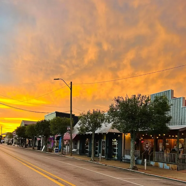 A street view during sunset with colorful clouds, featuring storefronts along the road lined with trees.