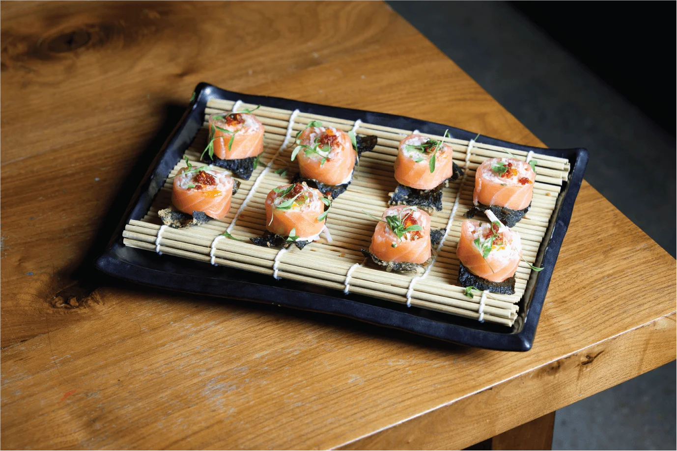 Salmon wrapped sushi bites topped with roe and microgreens, served on a bamboo mat atop a black rectangular plate.