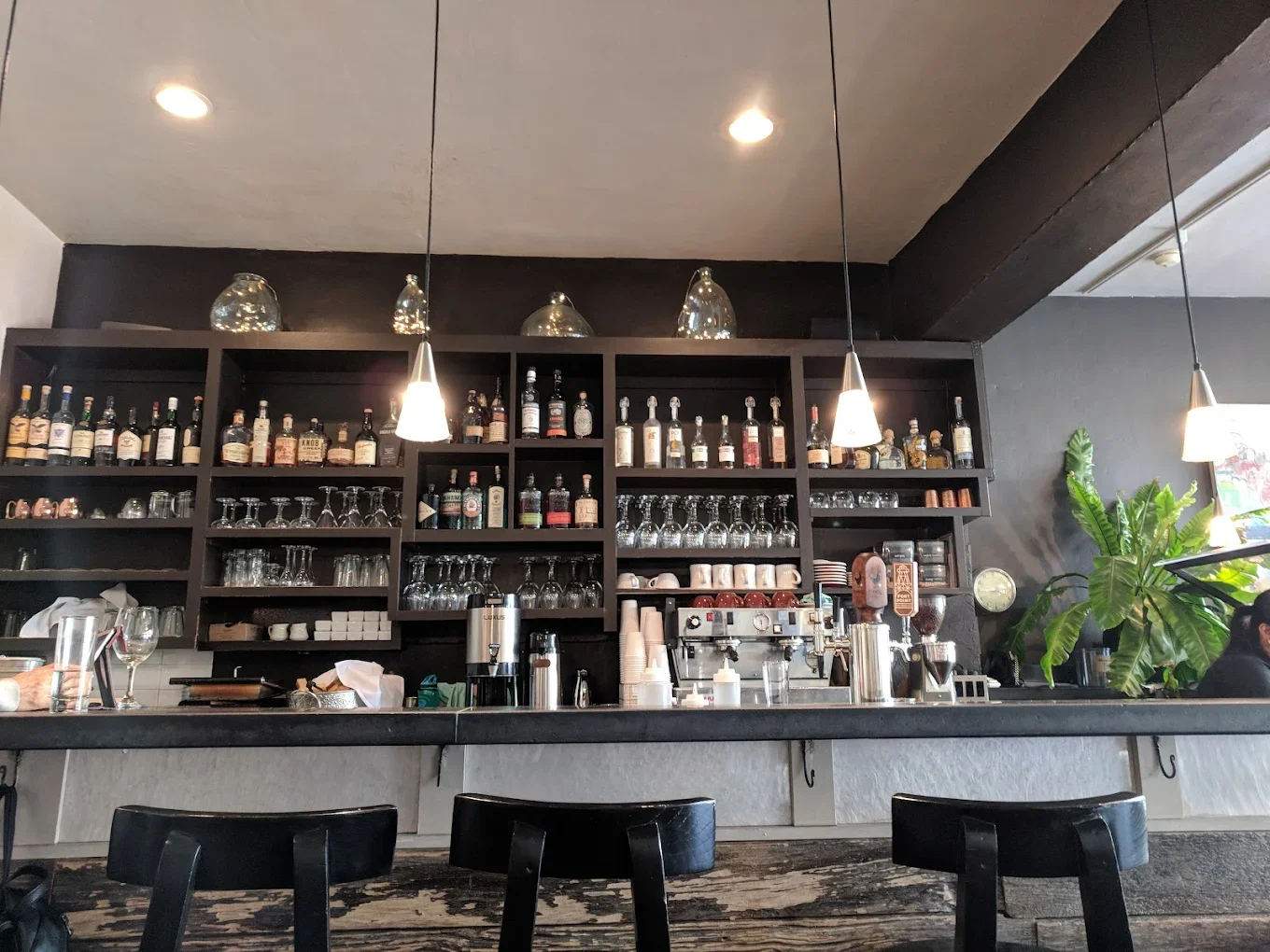 An interior view of a bar with a dark wooden shelf displaying various bottles of alcohol and glassware. Soft lighting highlights the bar area, creating a cozy atmosphere.