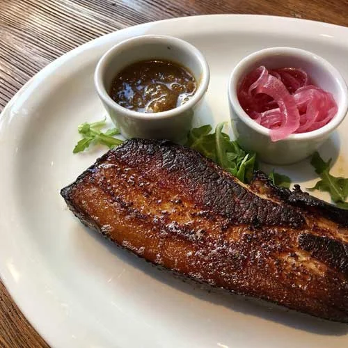 A white plate with seared pork, arugula, and two small cups, one with salsa and one with onions