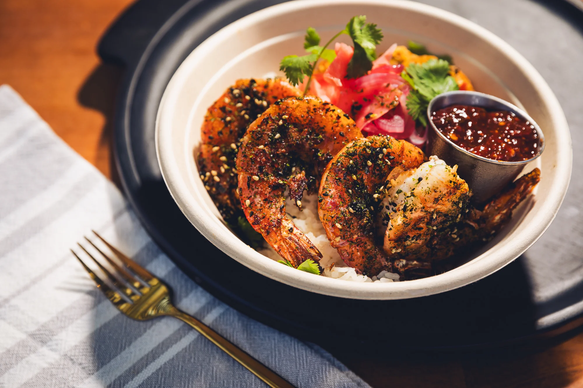 A bowl of seasoned shrimp served over rice, garnished with fresh herbs, pickled onions, and a small container of sauce.