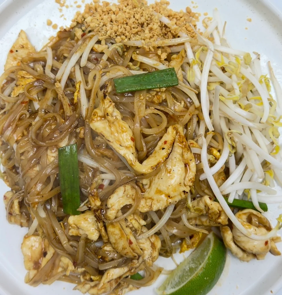 A plate of stir-fried noodles with chicken, bean sprouts, green onions, and crushed peanuts, garnished with a lime wedge.