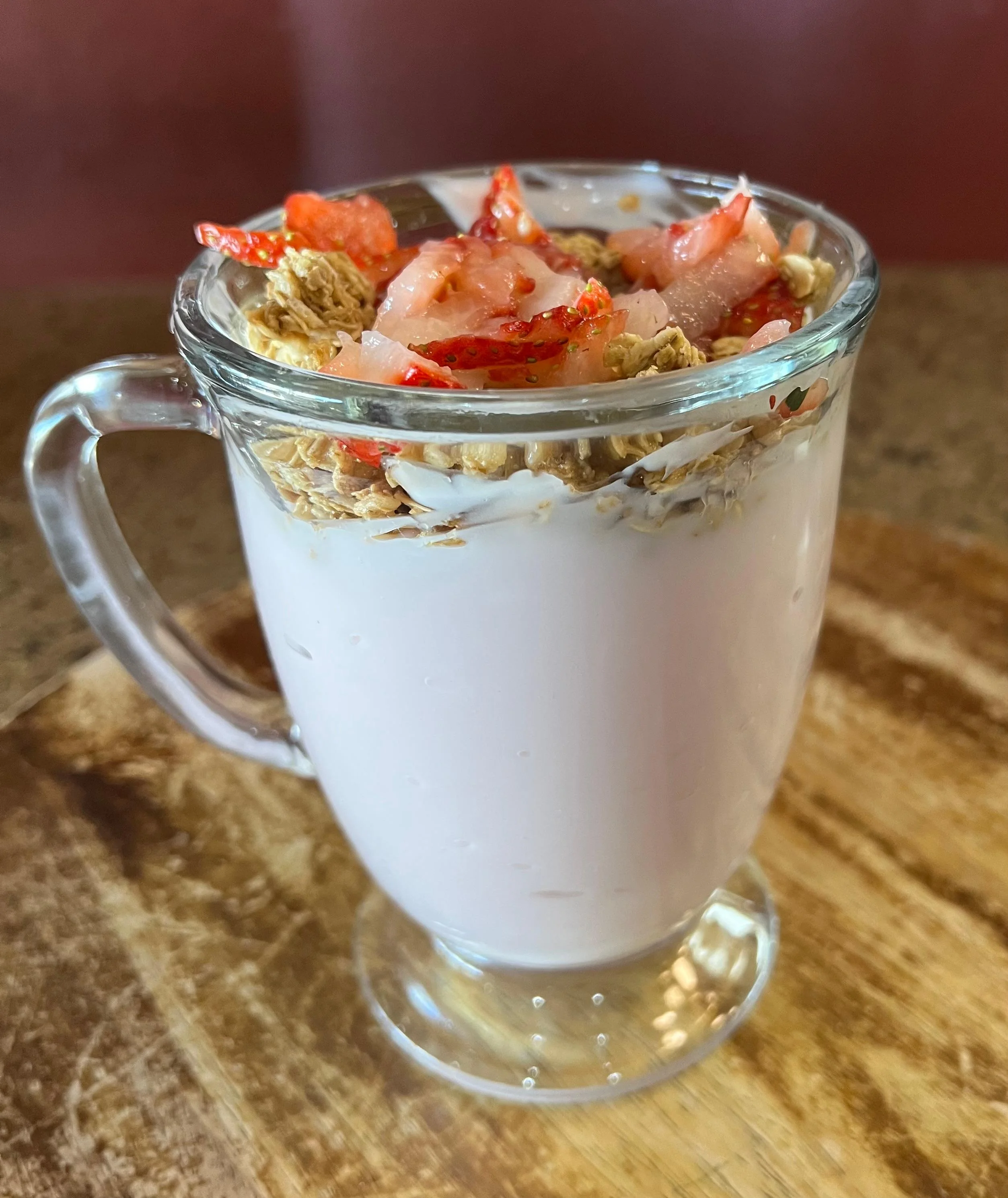 A glass parfait filled with yogurt, granola, and fresh strawberries, presented on a wooden surface.