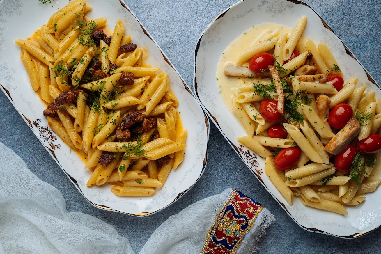 Two plates of pasta, one with meat and herbs, and the other with chicken and cherry tomatoes, garnished for presentation.